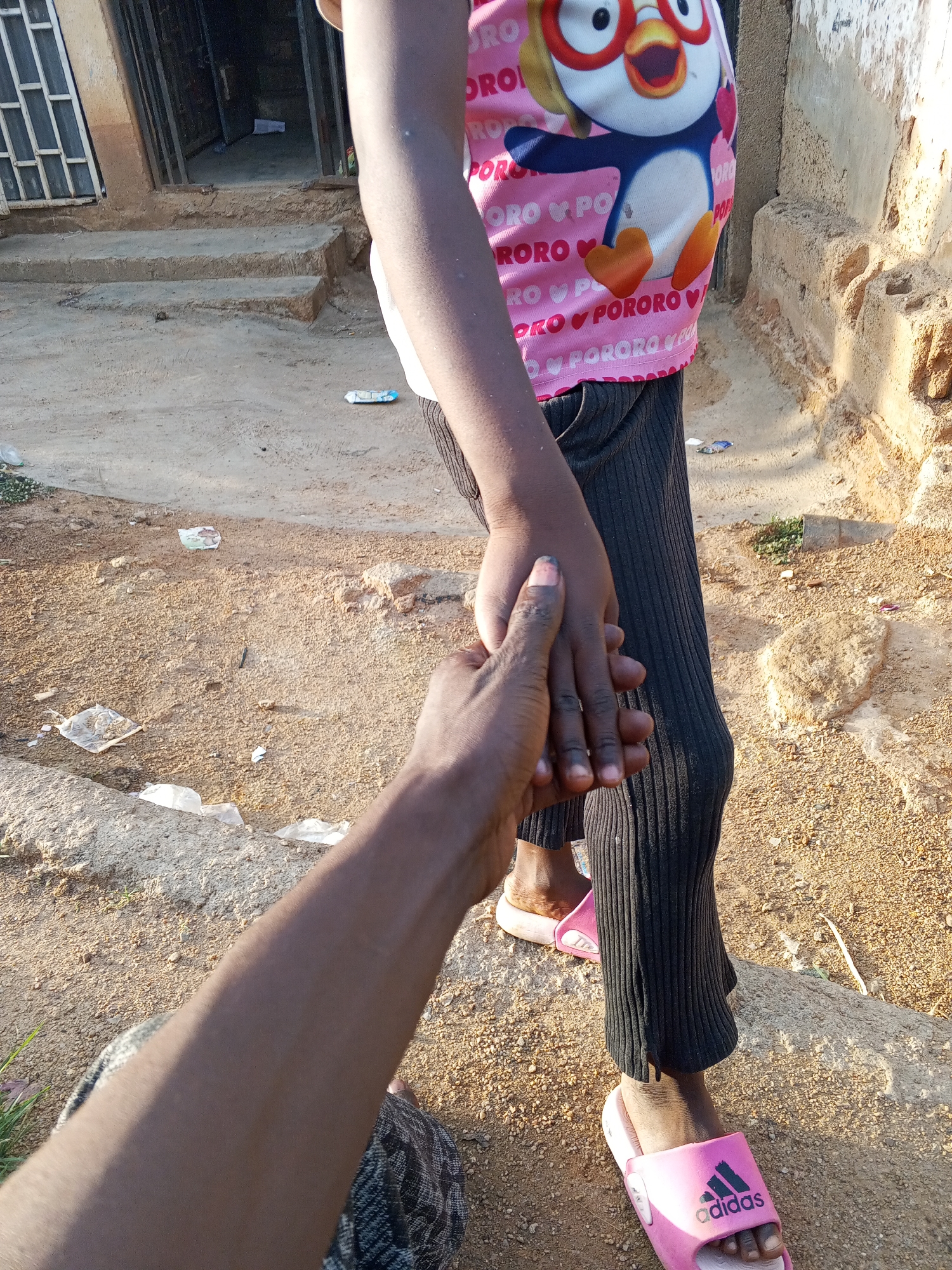 In Gwong, Nigeria, an individual extends their hand to a child wearing a pink Pororo-themed shirt and black leggings. The child is wearing pink Adidas sandals. They stand on a dirt ground scattered with debris. In the background are concrete steps leading to a building. The light suggests it is daytime.