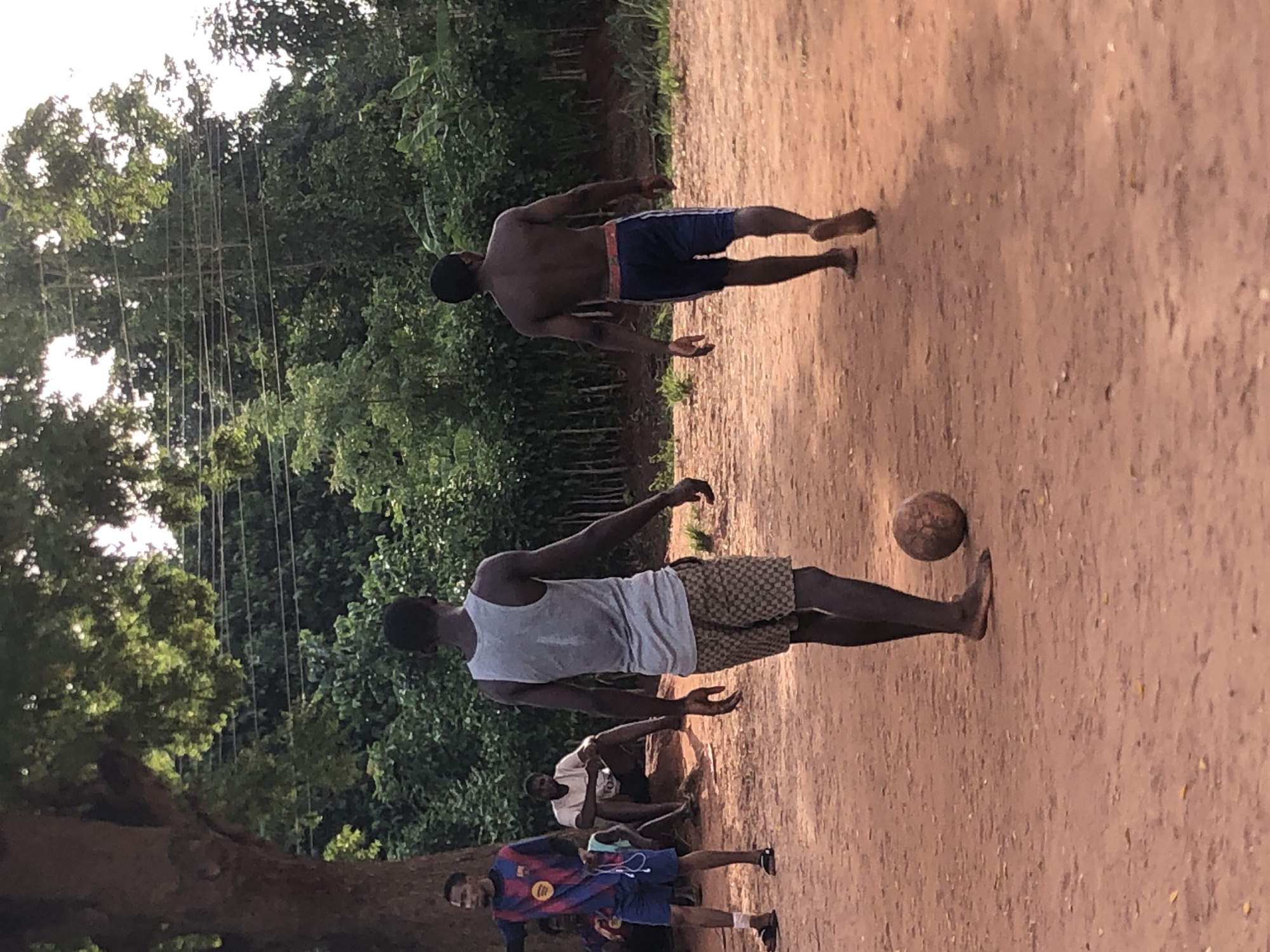This image captures a casual football (soccer) game or gathering in Ukwu-Abwa, Nigeria, set on a reddish-brown dirt field. The scene is outdoors, surrounded by dense, lush green trees and foliage under what appears to be a bright sky, suggesting daytime, possibly late afternoon.
In the foreground, a young man wearing a grey tank top and patterned shorts stands barefoot, facing away from the camera, with a worn, brown football near his feet. Behind him, slightly to the right, another man, shirtless and wearing blue shorts, is also barefoot and facing away, appearing to be part of the game. Further back and to the left, another individual in a blue and red FC Barcelona jersey and dark shorts, wearing sandals or shoes, is smiling towards the camera while holding what appears to be a phone or small device. Text "Barcelona" is visible on his jersey.
Other people are visible in the background; one man in a white t-shirt is seated, and others are partially obscured. The overall atmosphere suggests a relaxed, informal gathering for football. In the distant background, amidst the trees, faint vertical lines or structures, possibly clotheslines or a rudimentary netting system, are visible against the bright sky. The simplicity of the setting, including the dirt field and barefoot players, aligns with a rural or semi-urban environment.