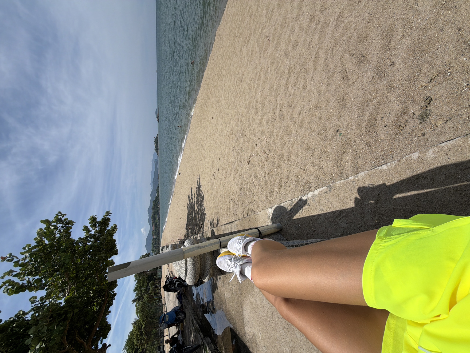 The image shows a first-person perspective of a person sitting on a concrete ledge at a beach in Nha Trang Ward, Vietnam. The person's legs, clad in bright neon yellow shorts and white athletic shoes with yellow accents, are positioned in the foreground. The beach itself is composed of light brown sand, with the turquoise ocean extending to the horizon under a partially cloudy sky. A few small islands are visible in the distance, as is a coastline with hills. To the left of the frame, a large tree with green leaves casts a shadow, and a concrete wall with stone accents runs along the beach. Several motorcycles and a blue scooter are parked on the concrete pathway behind the wall, where a person is seated. The overall scene depicts a relaxed day at the beach.