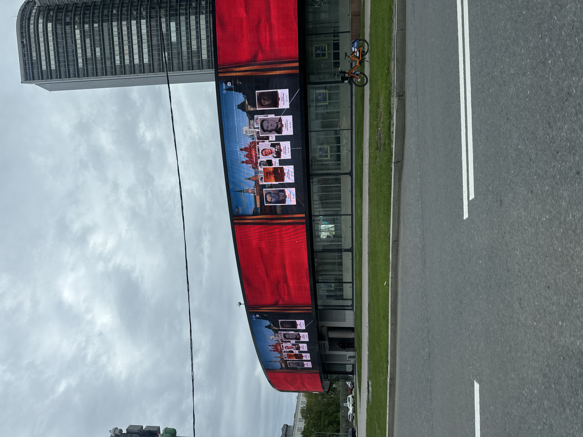 The image depicts a street scene in Moscow, Russia. A large digital billboard displays images of individuals over a backdrop of the Kremlin and other Moscow landmarks, with the Russian flag colors prominently featured. A tall skyscraper looms in the background, and a rental bicycle is parked along the sidewalk adjacent to the building housing the billboard. A multi-lane road with white lane markings is visible in the foreground. The sky is overcast.