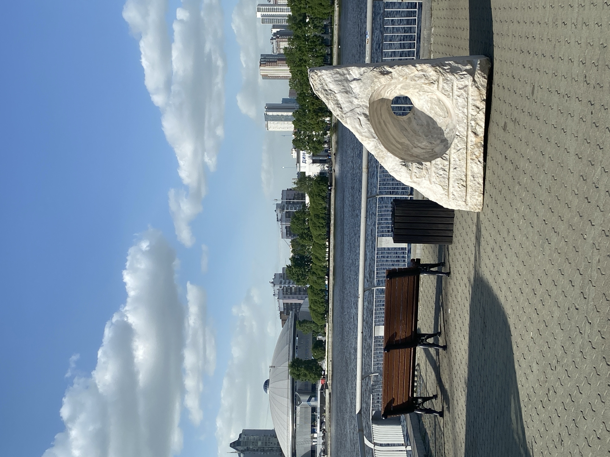 The image captures a scene from the city of Yekaterinburg, Russia.
The foreground shows a paved pathway next to a body of water, presumably the Iset River, judging by the presence of the Dinamo Metro Bridge and the Vysotsky business center in the background. A public park bench and a trash can are placed on the pathway. Beside them is a large, modern art sculpture made of light-colored stone or marble, with a hole carved through it, possibly intended for viewing the cityscape. The sculpture’s design suggests a contemporary aesthetic.
Across the river are buildings and trees, indicating an urban landscape. The presence of high-rise apartments suggests a densely populated area.
The sky is blue with white clouds, implying fair weather conditions. The image is taken from an elevated angle, suggesting the photographer was on a bridge or upper level. There are no people in the immediate view of the scene, focusing primarily on the cityscape and architectural elements.