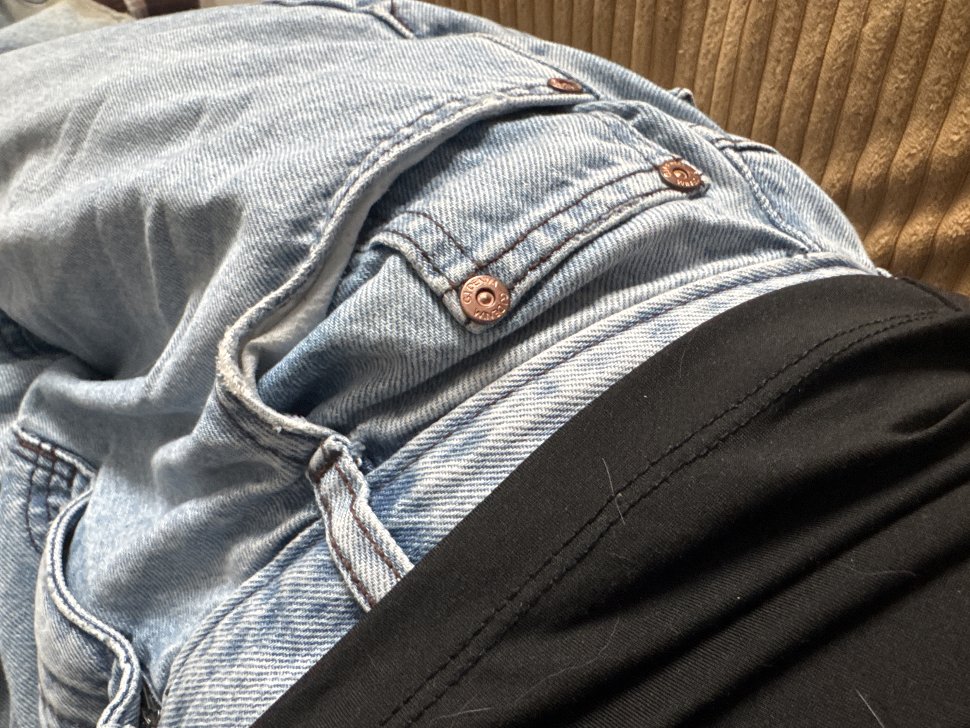 The image shows a close-up of a person wearing light blue denim jeans and a black top. The jeans have a small watch pocket with a metal button detail. The background is a textured, light brown surface. There is no information in the image to indicate it was taken in the city of Chaikovsky, Russia.