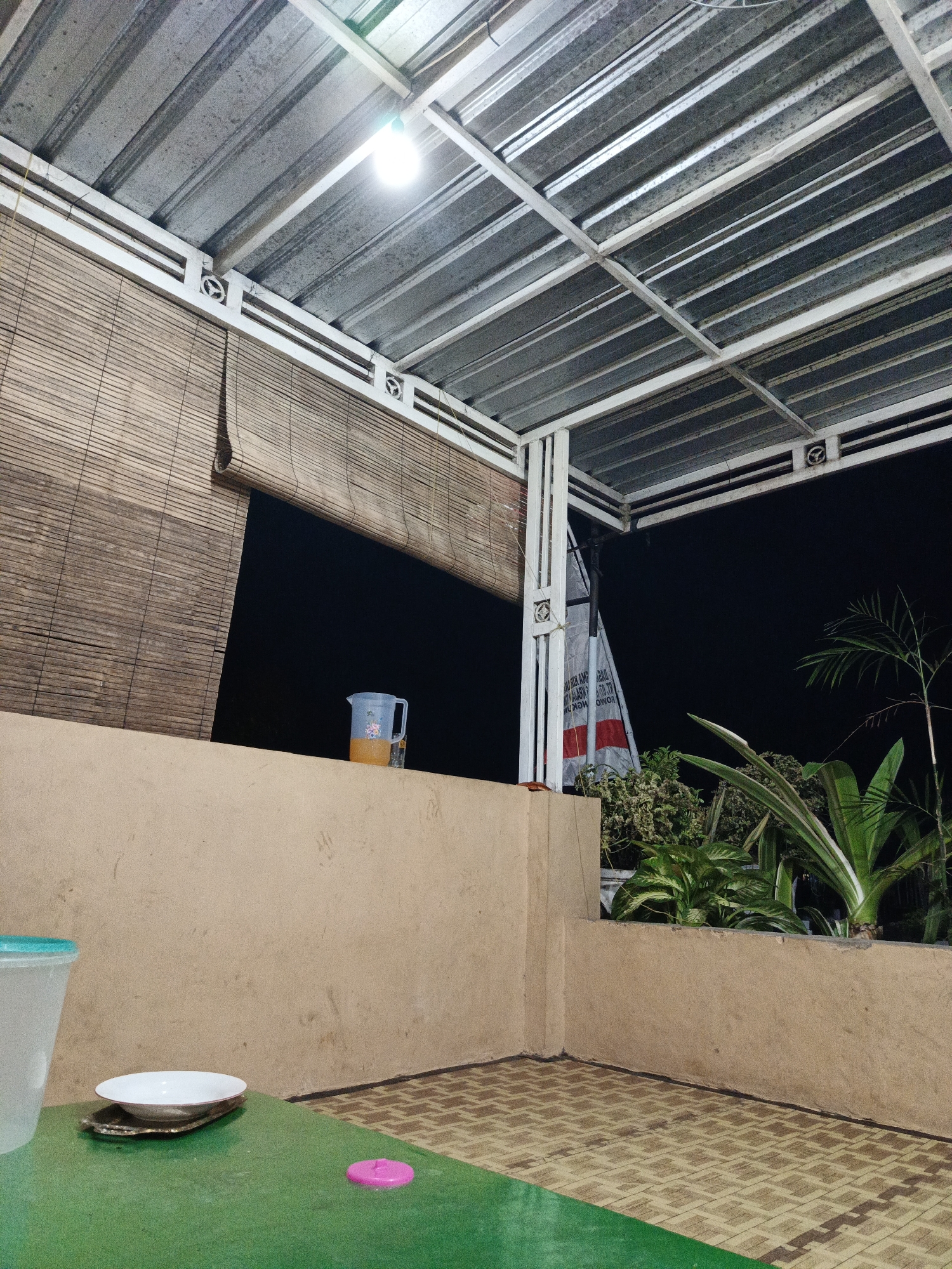 A nighttime view from a covered outdoor patio in Rowokangkung, Indonesia, shows a corrugated metal roof supported by white metal beams. A bamboo roller blind hangs partially drawn on the left side, revealing a dark background. On a tan-colored ledge, a clear plastic pitcher containing an amber liquid is placed next to a dark bottle. In the foreground, a green table has a white bowl on a decorative plate, and a bright pink object, possibly a lid or coaster, rests on the table's surface. The floor is tiled with a patterned design. Lush green plants are visible in the background, and a white banner with red and black text is partially obscured by a pillar on the right. A single light bulb illuminates the scene from the ceiling.