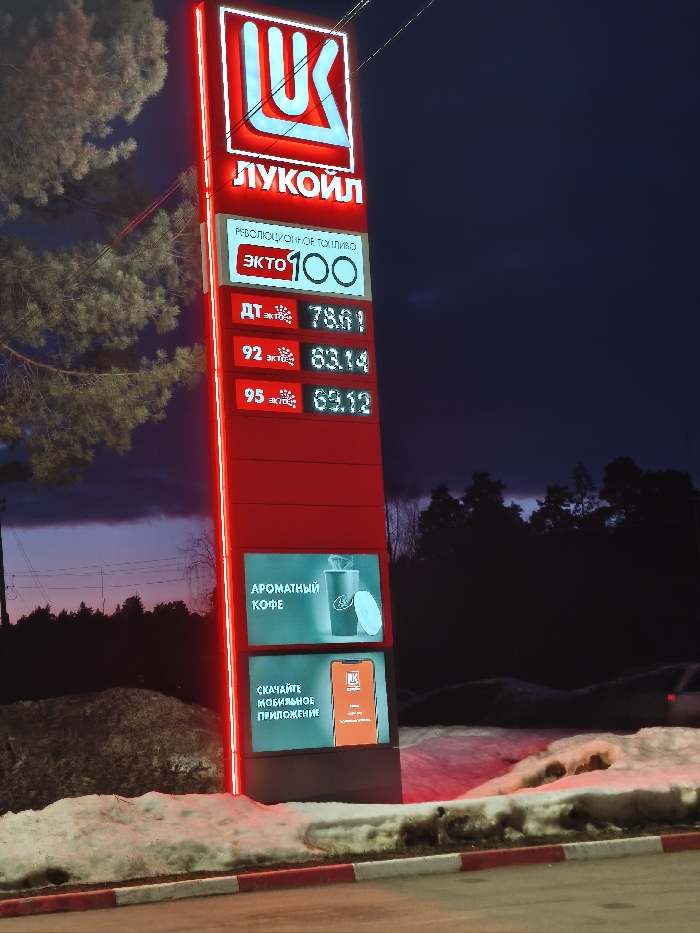 A brightly illuminated, vertical red LUKOIL gas station pylon sign stands prominently against a dark twilight sky in Sarapul, Russia. The sign displays the LUKOIL logo and name in white Cyrillic lettering at its apex, accented by red neon-style backlighting along its edges. Directly below, the phrase 'РЕВОЛЮЦИОННОЕ ТОПЛИВО' (REVOLUTIONARY FUEL) is presented in white, followed by 'ЭКТО 100' (EKTO 100) with 'ЭКТО' in red. Digital displays list current fuel prices: Diesel (ДТ) at 78.61, 92 octane at 63.14, and 95 octane at 69.12, each preceded by a red asterisk-like symbol. The lower section of the pylon features two integrated digital screens: the upper advertises 'АРОМАТНЫЙ КОФЕ' (AROMATIC COFFEE) with an image of a LUKOIL-branded cup, and the lower promotes 'СКАЧАЙТЕ МОБИЛЬНОЕ ПРИЛОЖЕНИЕ' (DOWNLOAD MOBILE APPLICATION) with an image of a smartphone displaying the app. Patches of dirty snow and brown earth cover the ground in the immediate foreground, bordering a red and white striped curb. A large conifer tree is partially visible to the left, illuminated by the sign, while a dark treeline frames the distant horizon on the right. No individuals, overt actions, or interactions are visible within the frame.