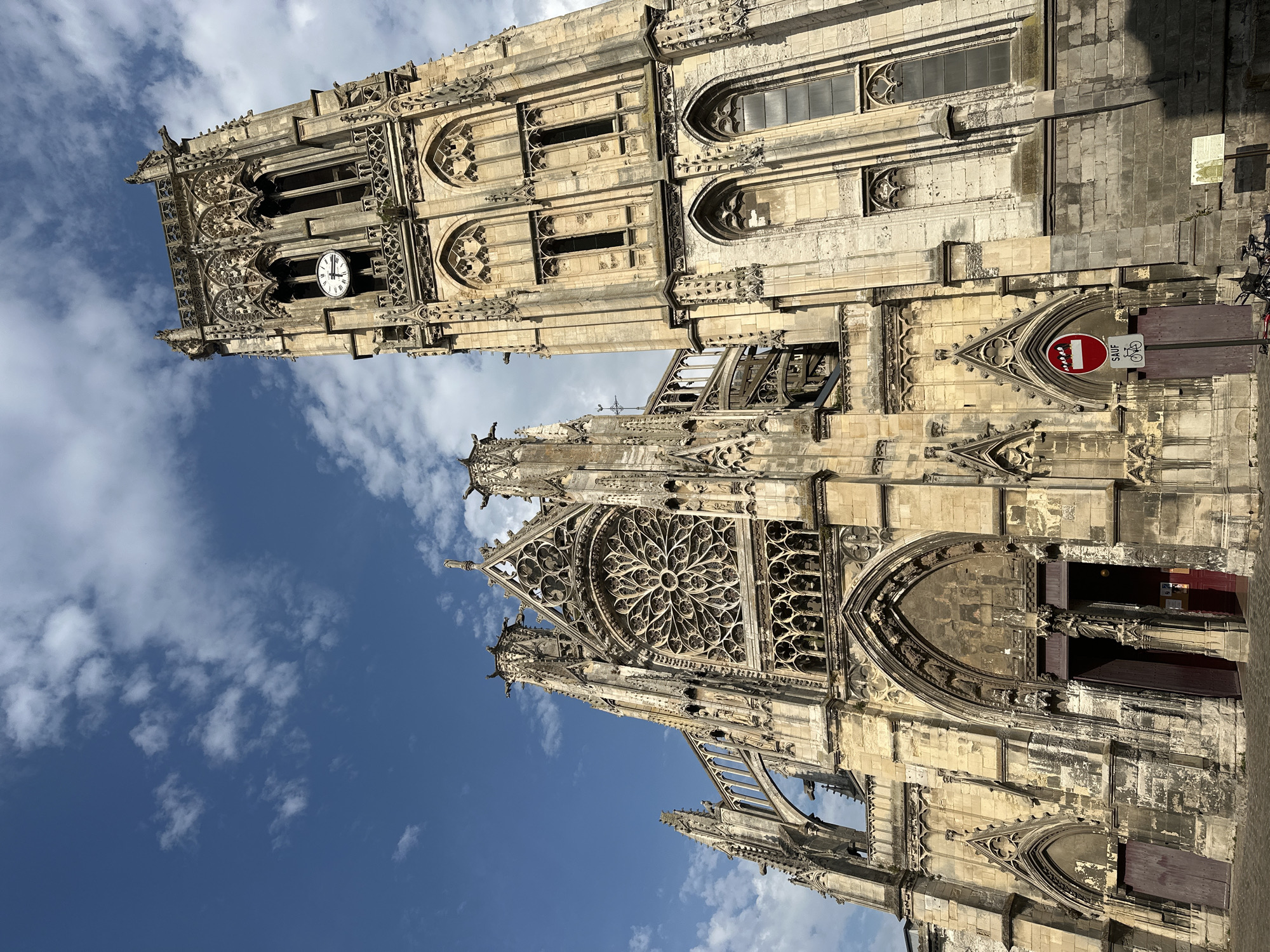 The image captures a low-angle view of the Saint-Jacques Church in Dieppe, France, under a partially cloudy blue sky. The architecture is Gothic, characterized by intricate stone carvings, pointed arches, and a large rose window on the facade. To the right, a tall bell tower with a clock face is prominent. A circular red sign with a white border, depicting a person walking and a bicycle, is affixed to a pole in front of the church. The ground in front of the building appears to be paved with cobblestones. No people are visible within the frame.