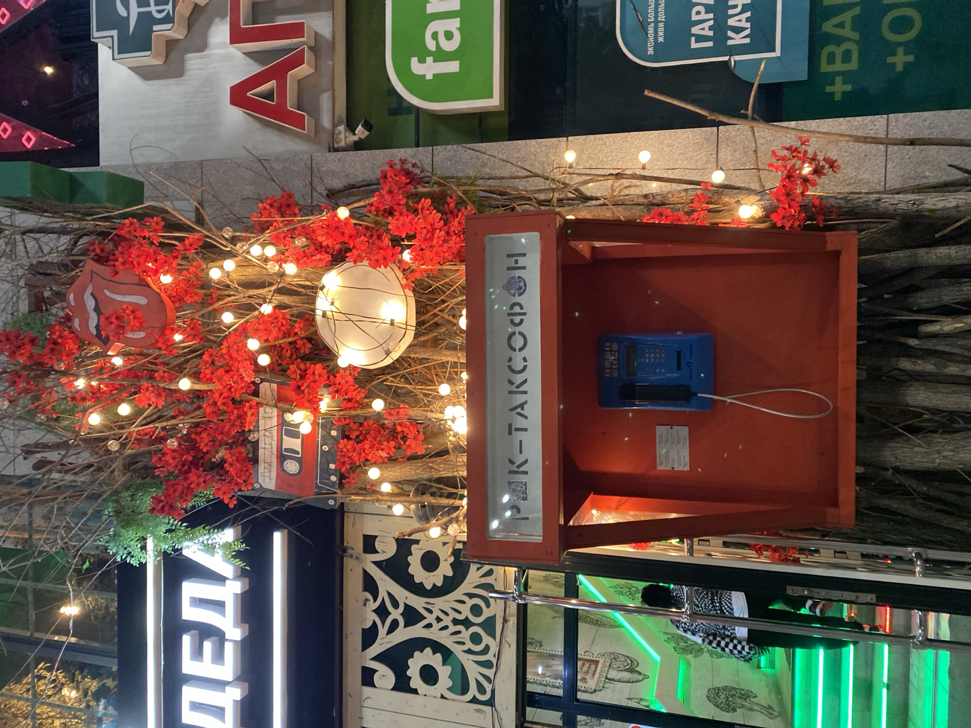 The image captures a unique urban scene likely located in Yekaterinburg, Russia, given the Cyrillic script and architectural style.
A red phone booth, labeled "PK-TAКСОФОН" is adorned with branches, red flowers, and miniature lights. A blue telephone is mounted within the box. To the booth's left, a sign "ДЕДА" illuminates vertically. Other signs with text, including "far", "ΓΑΡΙ", and "+BA", are visible.
In the background is a light colored stone wall. Additionally, portions of a building's exterior are seen, featuring glass panels and decorative patterns.
The setting appears to be outdoors, possibly at night. The presence of lights suggests an evening scene in an urban environment. There is a reflection on the glass of the sign. A person wearing black pants with a black and white patterned shirt can be seen reflected.