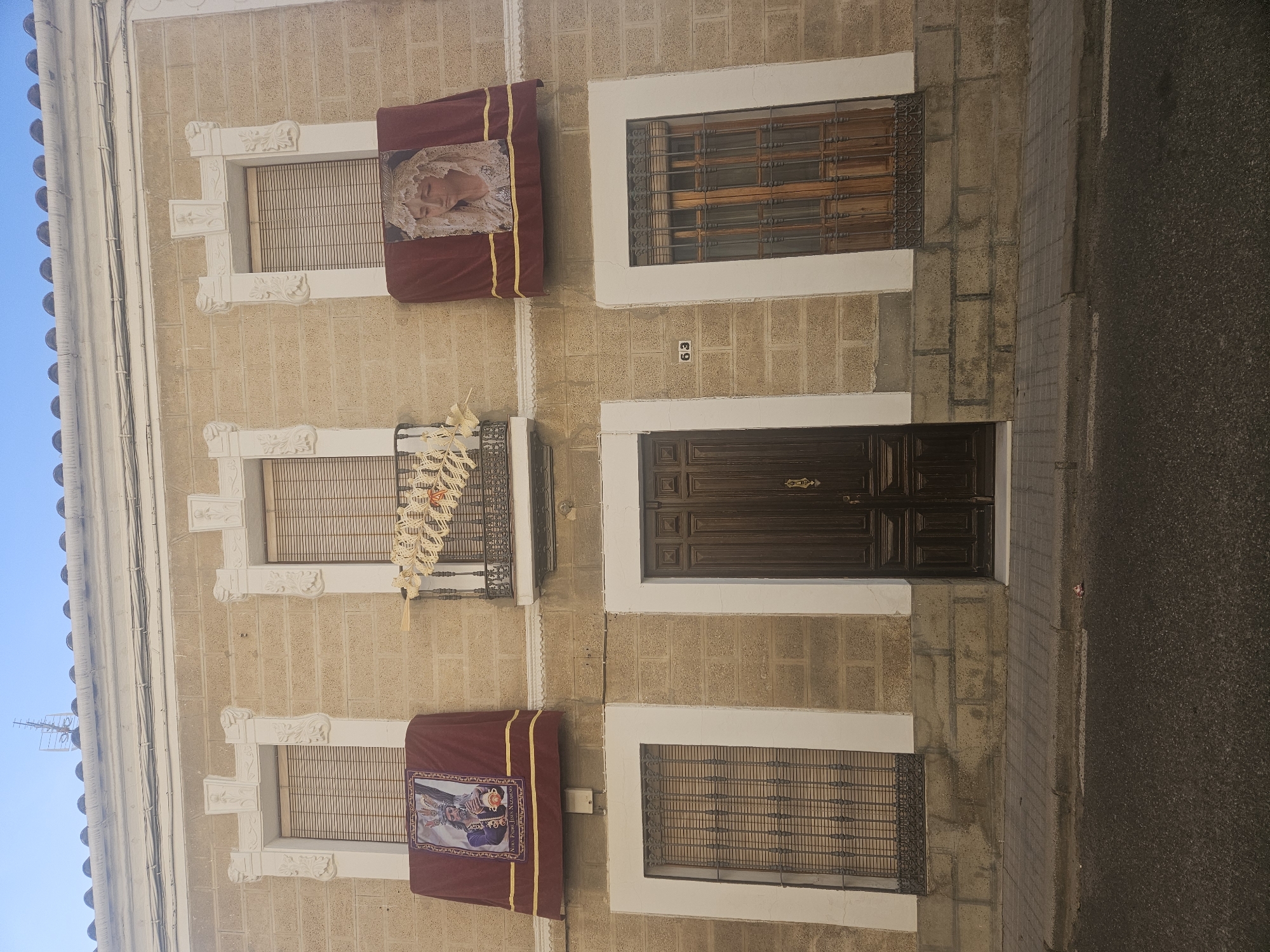 A vertically oriented exterior shot depicts a building facade in Pedro Abad, Spain. The facade consists of light brown stonework, with three stories visible. The windows on the left side of the facade are adorned with ornate white frames. The top and bottom windows on the left side of the facade have burgundy-colored drapery with a gold border. The top window features an image of a woman in a white veil. The bottom window features an image of a man in traditional clothing. The middle window on the left side features a metal balcony and is decorated with white fabric. The windows on the right side of the facade have white frames and metal bars. A dark brown wooden door is situated in the center of the facade. The number "69" is placed above the door. A section of the street is visible on the right side of the image. The sky is visible on the left.