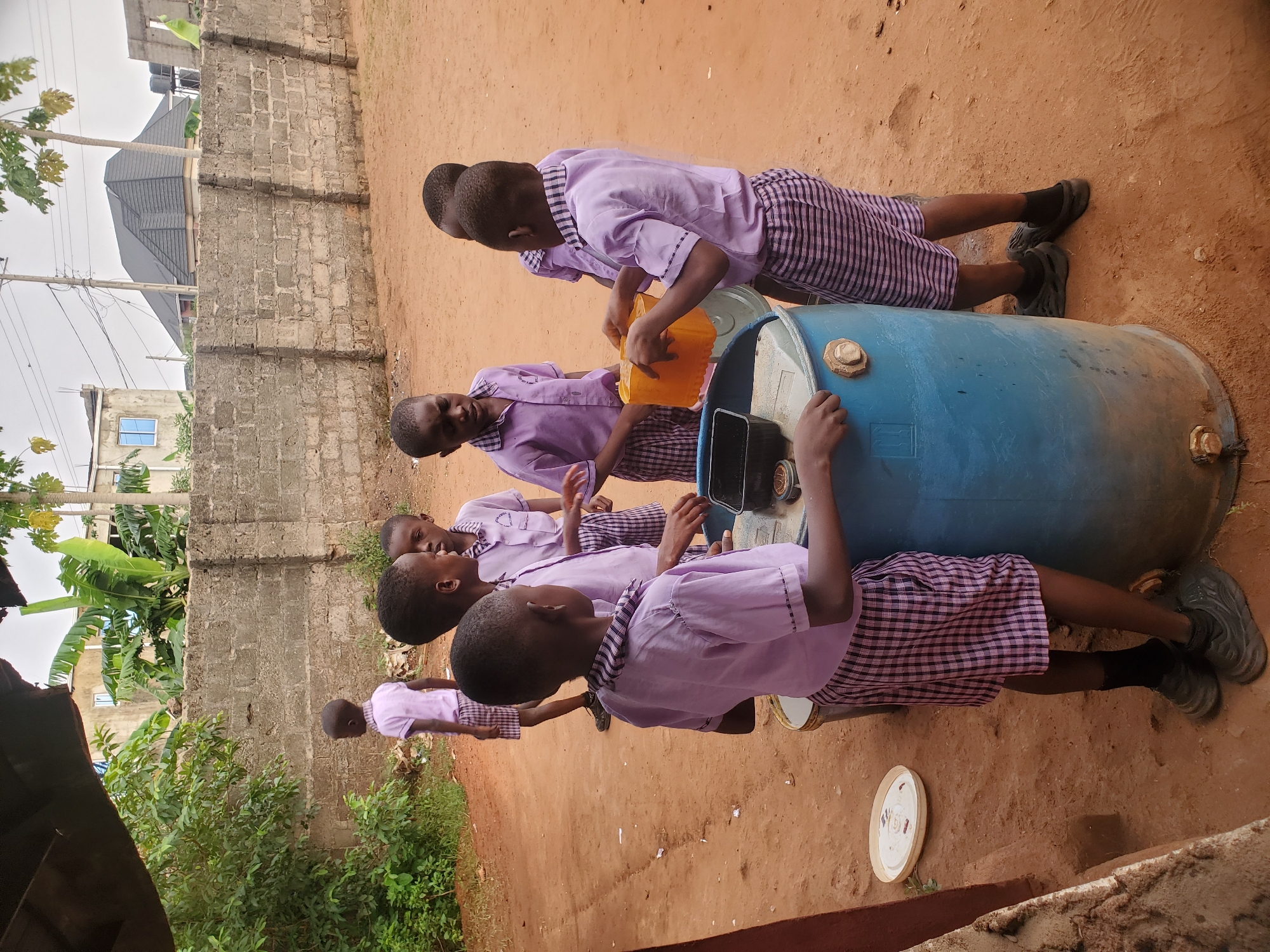 In Ukwu-Abwa, Nigeria, several children in purple school uniforms are gathered around a large blue barrel. One child is pouring water from a yellow container into the barrel. A black object, possibly a phone or small tablet, is placed on top of the barrel. The ground is reddish-brown. A brick wall is visible in the background, along with greenery and parts of buildings. The scene appears to be outdoors.