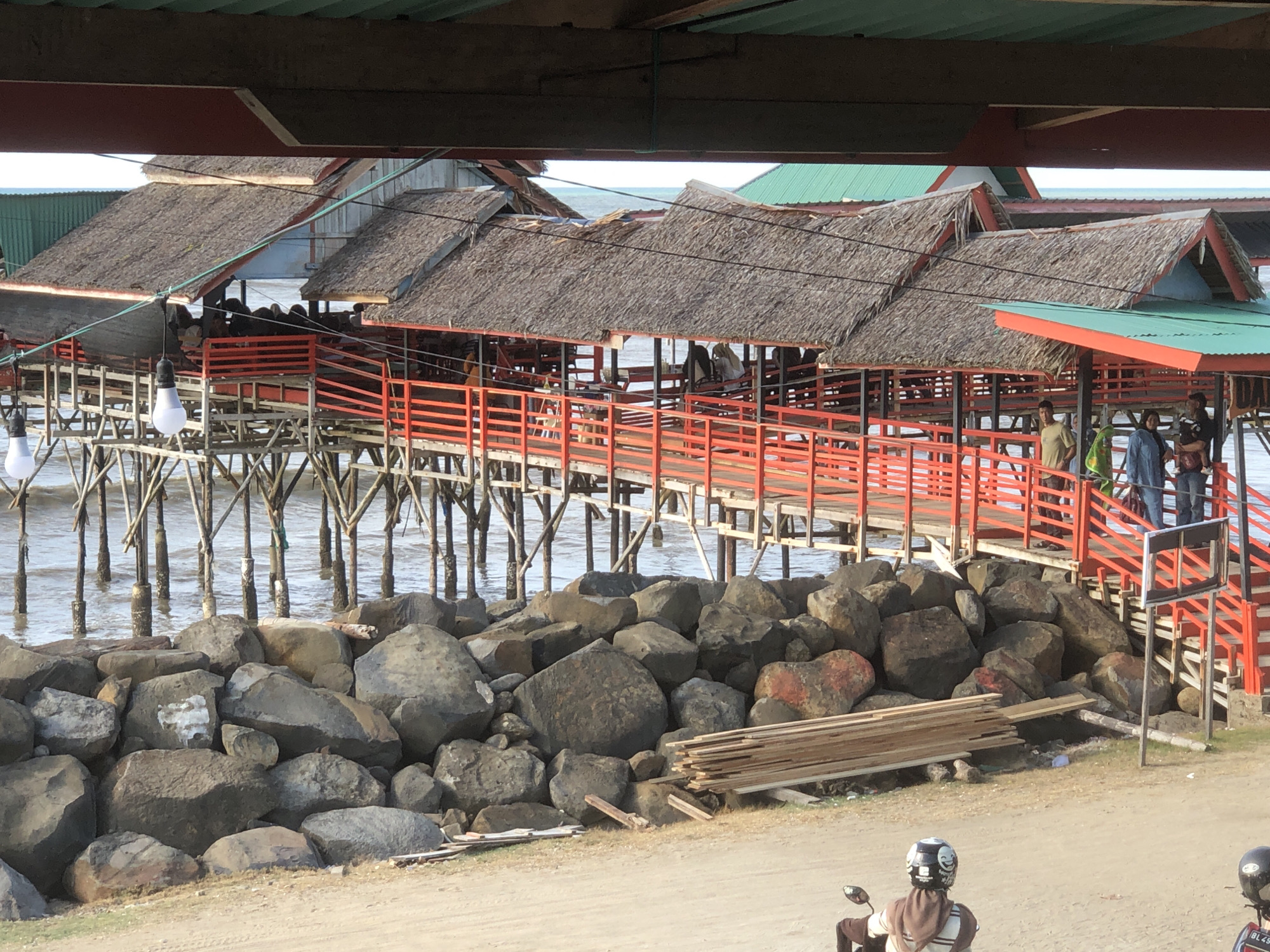 The image shows a waterfront view in Lancok, Indonesia, featuring a series of stilt-supported structures extending over the water. The structures have thatched roofs and red railings, appearing to be connected by walkways. Several people are present on the structures. Below the structures, there's a barrier of large rocks along the shore. In the foreground, a dirt road is visible, with a motorcycle parked. Wooden planks are placed adjacent to the rocks.