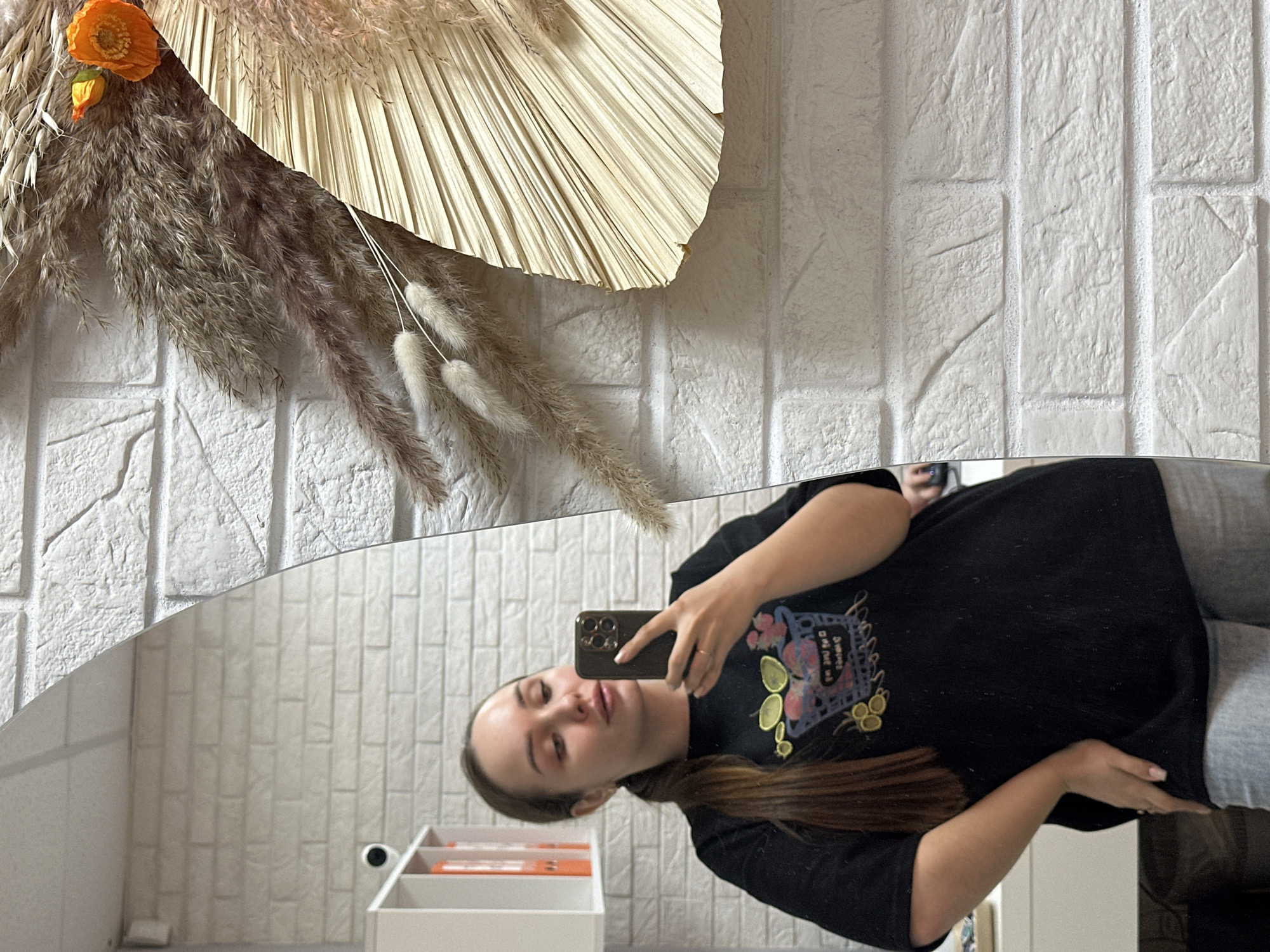A young woman takes a selfie in a mirror. The mirror is mounted on a white brick wall decorated with dried flowers and leaves. In the mirror's reflection are a white shelf containing orange items and another section of the white brick wall. The woman is wearing a black t-shirt with a colorful graphic. The city of Chaikovsky, Russia, cannot be determined from the image.