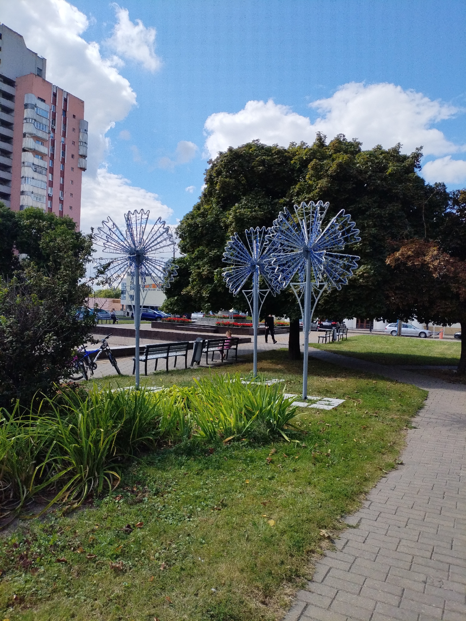 This is a daylight photograph taken outdoors on a street in Mahilyow, Belarus. The scene depicts a sidewalk on the right, made of gray brick pavers arranged in a herringbone pattern. Adjacent to the sidewalk is a well-maintained grassy area with a few scattered fallen leaves. In the foreground, tall, green grassy plants are growing. Two decorative light structures resembling large, stylized dandelions with blue-white lights are prominently featured in the mid-ground, positioned in the grassy area. Behind these, several dark green trees create a backdrop, with one tree on the right exhibiting brown foliage. Further back, a row of dark metal benches is visible. A blue bicycle is parked near the left edge of the frame, leaning against what appears to be a low, dark-colored wall or planter. Beyond the benches and trees, a building with a light pink facade and multiple balconies is visible on the left. Other cars and buildings are present in the background, along with a blue sky with scattered white clouds. A young child with pink clothing is sitting on one of the benches, facing away from the camera. Another person, dressed in dark clothing, is walking away from the camera on the sidewalk.
