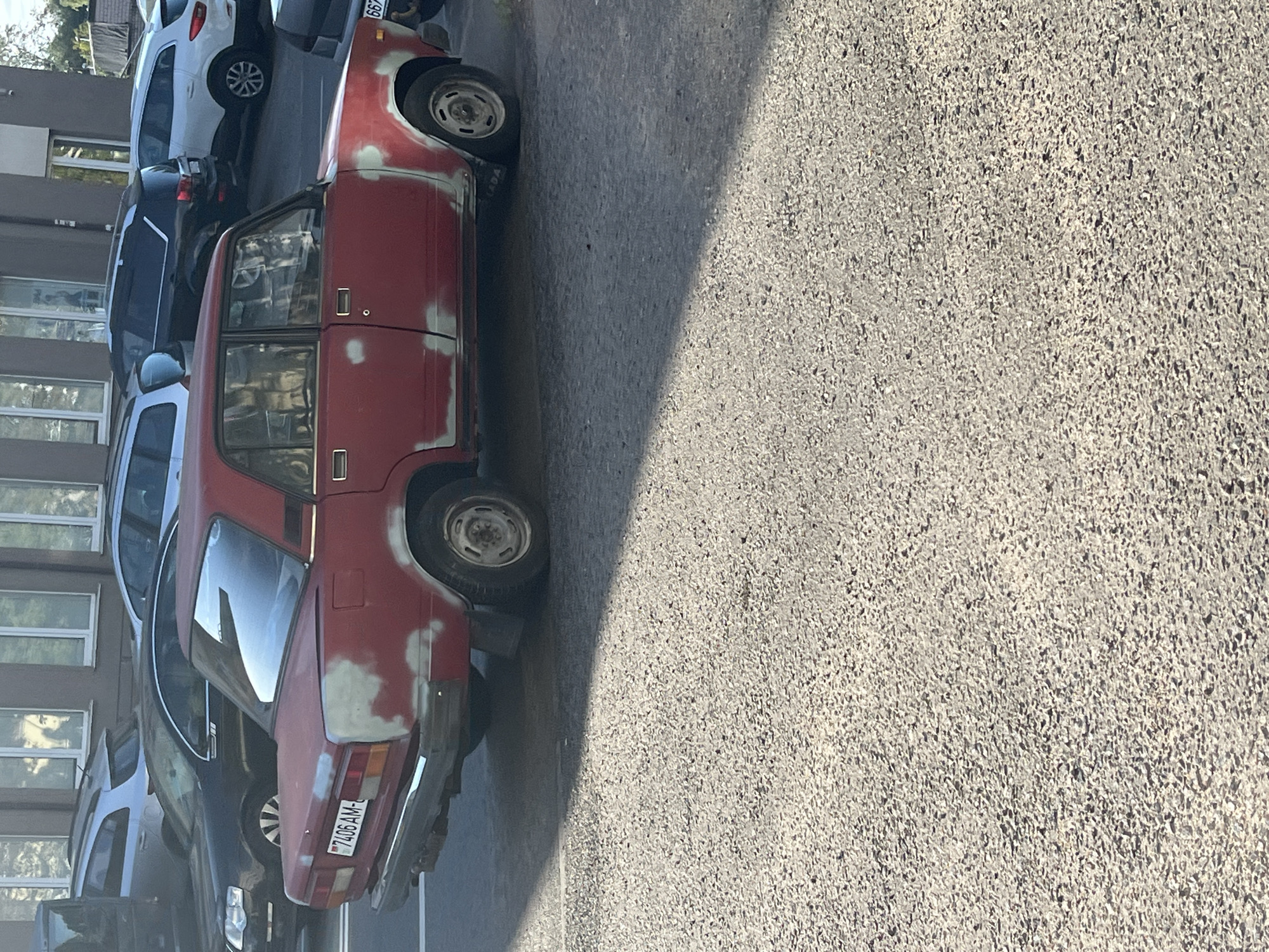 A red Lada car, model 2107, with visible primer patches on its body, is parked on a paved street in Minsk, Belarus. The car has a license plate reading "7406 AM-4". In the background, several other vehicles are parked, including a black Volkswagen Touareg SUV, a white Renault Duster SUV, and a dark grey sedan. The street appears to be in a residential or commercial area, with a building visible in the background with windows reflecting the sky. The scene is illuminated by sunlight, casting shadows on the pavement.
