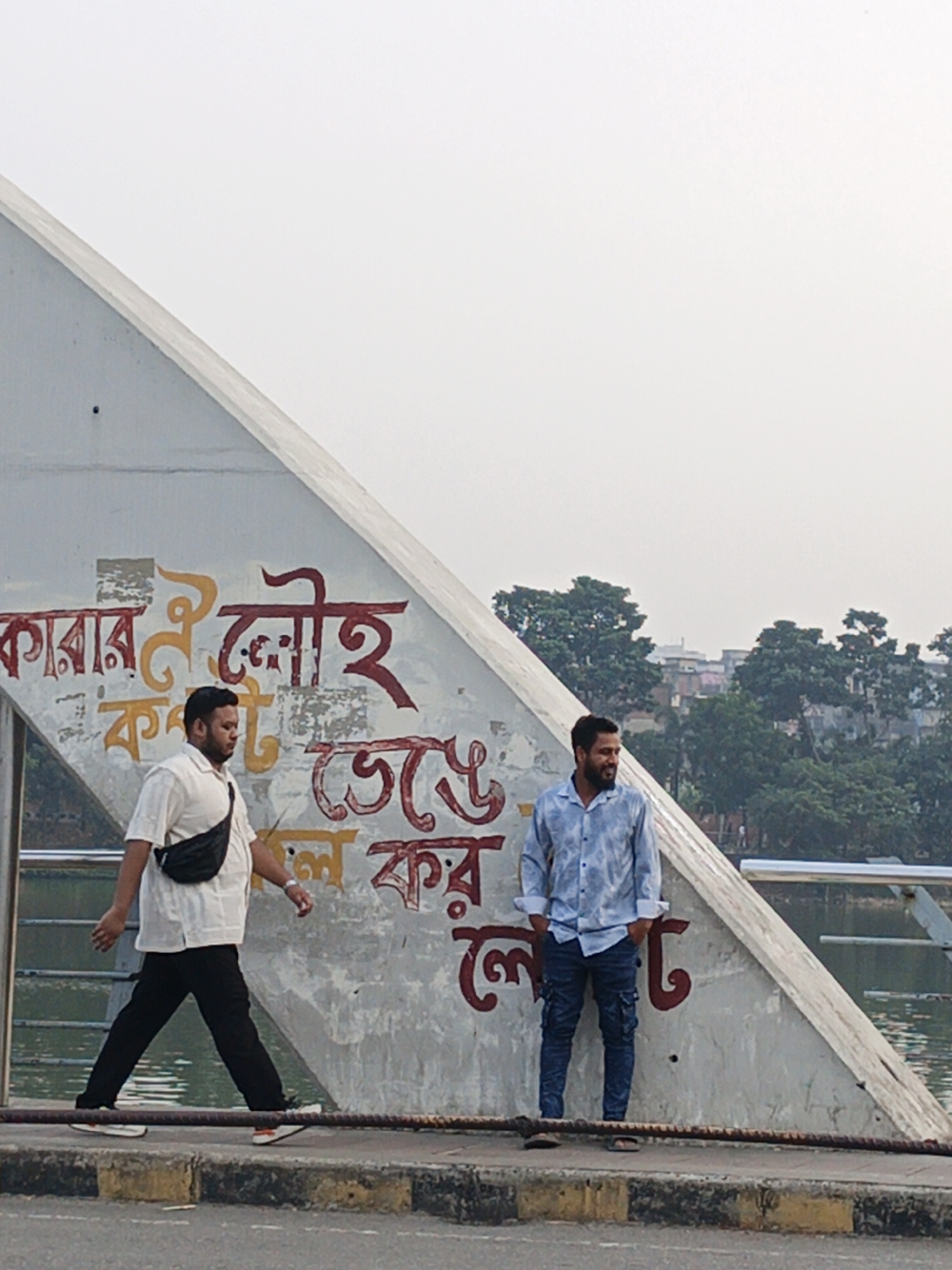 Two men are on a bridge in Dhaka, Bangladesh. The bridge has a large concrete arch with text written in Bengali on its surface. The man on the left is walking, wearing a white shirt, black pants, and a black fanny pack. The man on the right is standing, wearing a light blue patterned shirt and dark jeans. A body of water is visible behind the bridge. The background shows urban buildings and trees. The sky is overcast.