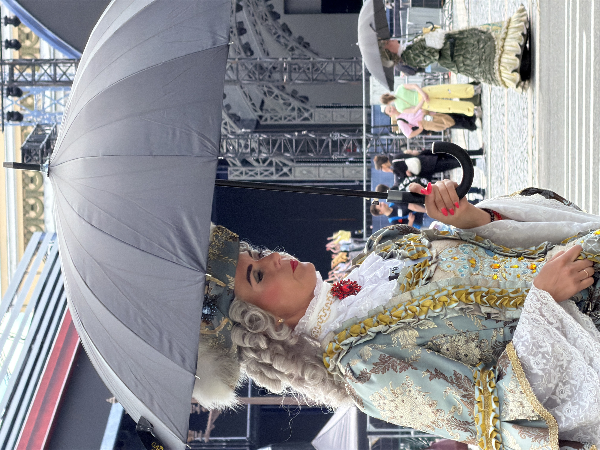 In Saint Petersburg, Russia, a person in an elaborate 18th-century costume, complete with a powdered wig, a richly embroidered pale blue and gold brocade jacket, and a white ruffled jabot adorned with a red jeweled brooch, is holding a large gray umbrella. The person's makeup is dramatic, with rouged cheeks and red lipstick. They are partially obscured by the umbrella, which is angled upwards.
In the background, a crowd of people are visible, some in modern clothing, others in historical dress, suggesting a reenactment or festival. One person in the background wears a green patterned dress and a ruff, and another in yellow trousers. Behind them, large metal structures, possibly staging or scaffolding for a performance, are visible. The lighting suggests it is daytime, and the weather is fair, as umbrellas are being used. The overall scene implies a public event with a historical theme taking place in an urban setting. There is no visible text in the image.