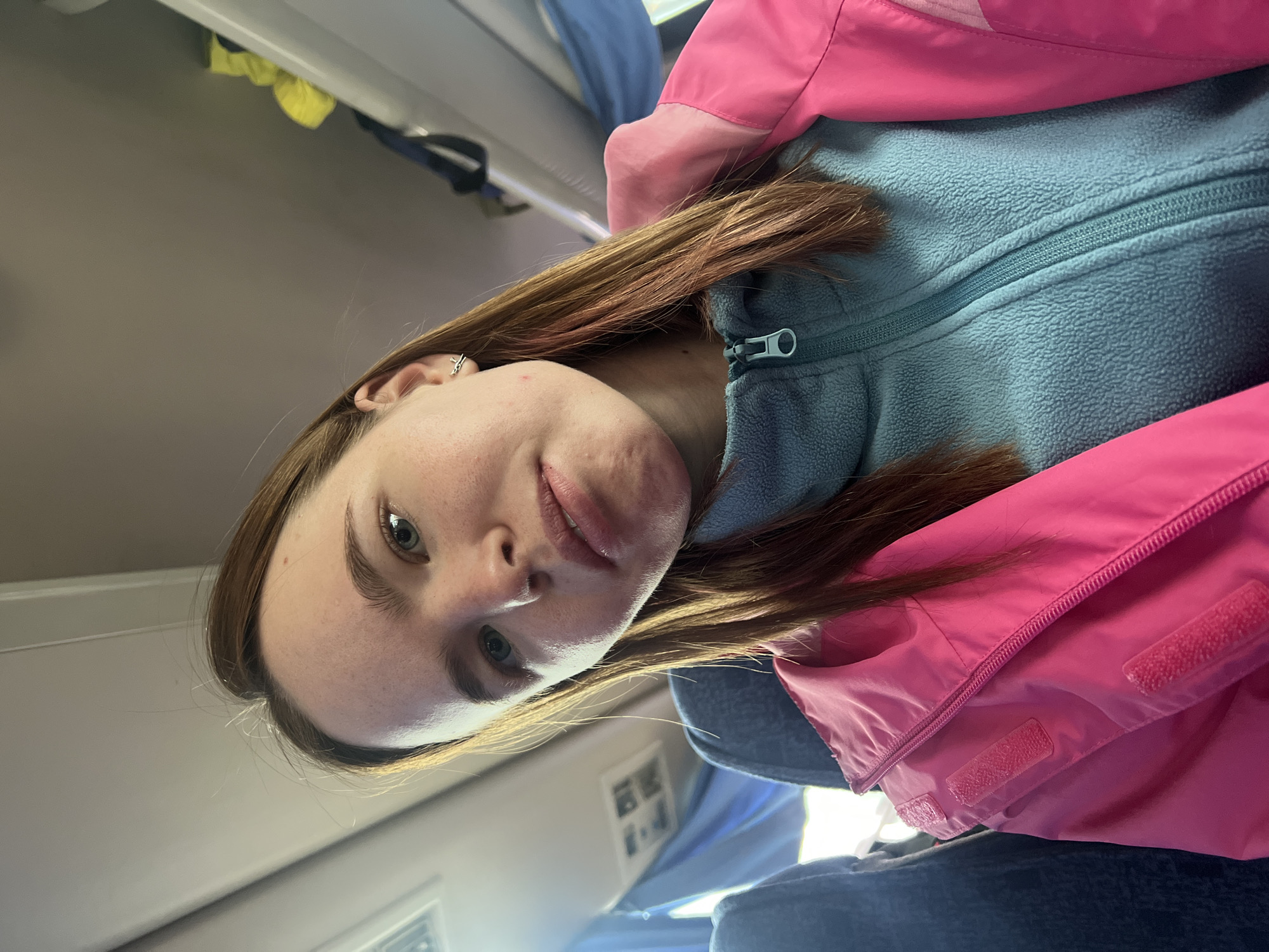 A young woman with light brown hair is wearing a light blue fleece jacket with a zipper and a bright pink waterproof jacket. She is positioned in a seated location, likely on a train or bus, as suggested by the overhead compartment and seat visible in the background. The location is Krylovo, Russia.