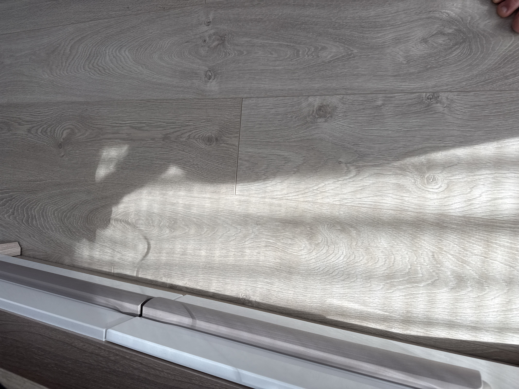 The image shows a close-up of a light gray wood-grained floor in what appears to be an interior setting. The floor is made up of planks, visible from the seams running across the surface. There are shadows cast across the floor, suggesting natural light from a window or opening. In the lower portion of the image, there is a layered structure consisting of what seem to be parts of a built-in element or furniture piece. The lower layer is a darker wood-grain, and on top of that, there are white and gray sections with a layered appearance. No people are visible in the majority of the image, except for a small portion of a hand in the upper-right corner. There is no indication of this image being located in Lovech, Bulgaria.