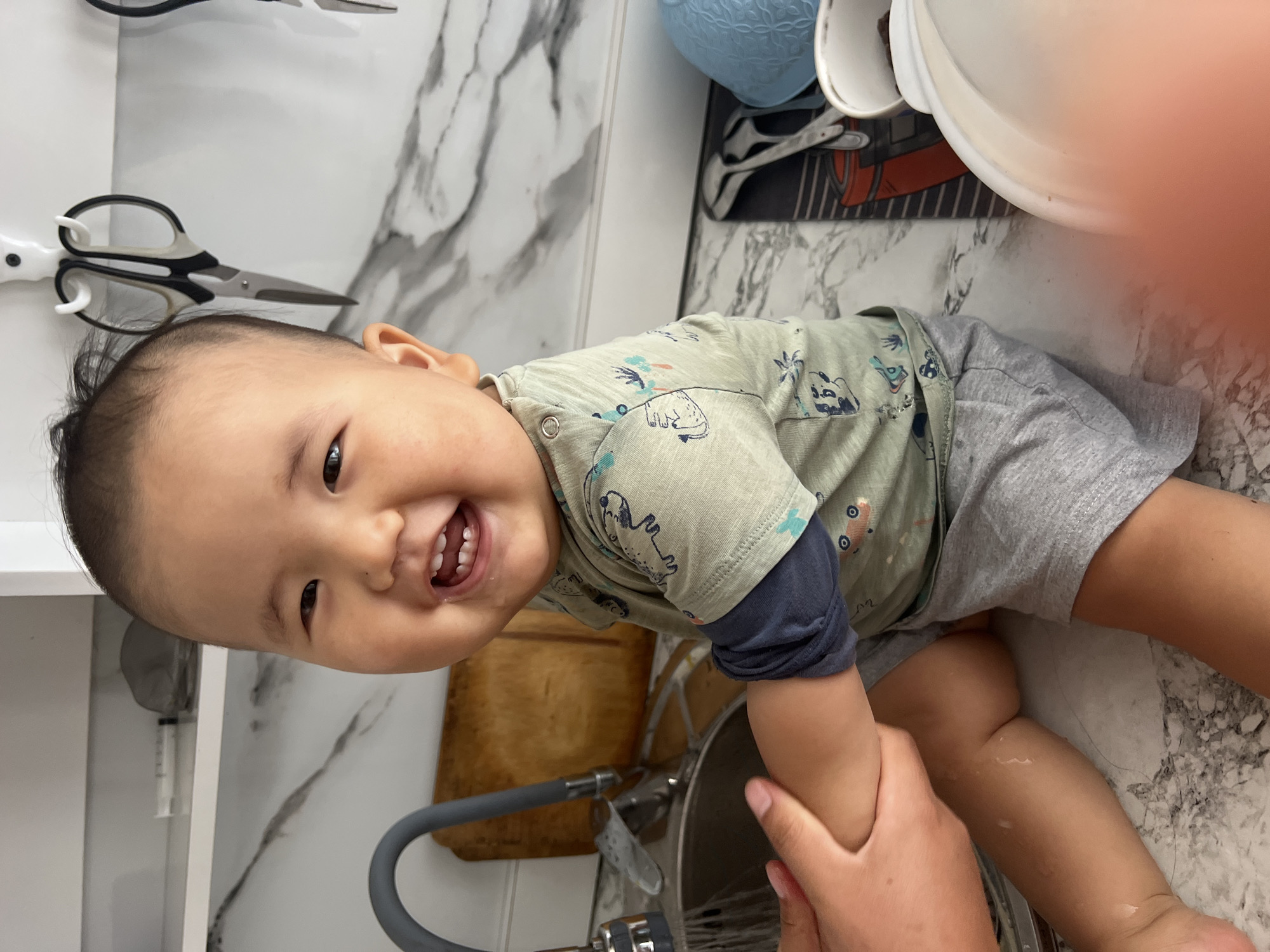 A young East Asian child, appearing to be between 6-12 months old, is seated on a countertop with a marble-patterned surface. The child is wearing a light green short-sleeved shirt with a dark blue long-sleeved shirt underneath, and grey shorts. The child has a joyful expression, with mouth open and teeth visible. The child's left arm is being held by an adult's hands. To the left of the child, a stainless steel faucet with a grey handle is visible, extending over a sink. Behind the child, a wooden cutting board is propped up against a white shelf unit. On the shelf, there are various items including what appears to be a syringe and a small grey object. To the right of the child, on the countertop, there is a black mat with utensils, a blue bowl, and a white bowl. The background consists of a white wall with a marble-patterned backsplash. The scene appears to be a domestic kitchen environment. The location is not discernible from the image itself and therefore cannot be placed in Kara-Dıykan, Kyrgyzstan.