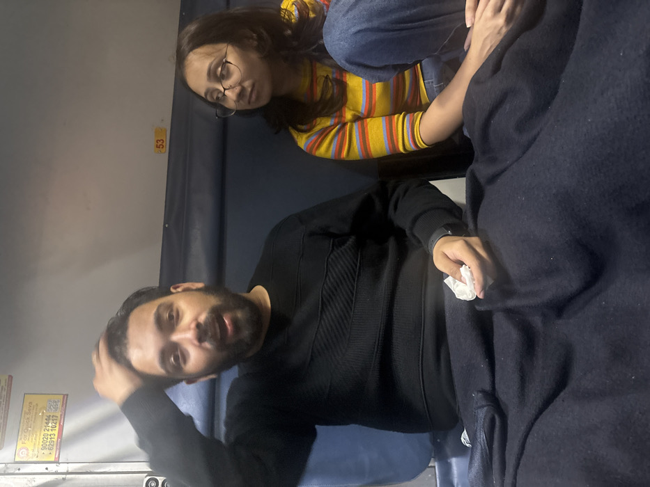 The scene depicts the interior of a train coach in India, traveling to or from Sitarampur. Two individuals are seated on a blue upholstered berth. A male, with a beard and dark hair, wearing a black long-sleeved sweater with textured horizontal patterns, occupies the left side of the frame. His mouth is open, suggesting speech, and his right hand is raised, touching the top of his head. His left hand, resting on a dark blue blanket covering his lap, holds a crumpled white tissue. He wears a dark-colored watch on his left wrist. To his right, a female with long dark hair and glasses is seated, looking towards her right, away from the male. She wears a yellow long-sleeved striped shirt with red, blue, and light blue stripes, and blue jeans. Both are partially covered by the dark blue blanket. In the background, a light grey wall with a blue rail runs horizontally. A yellow sticker with Hindi text and contact numbers (e.g., "90020 21444", "62913 10217") is visible on the upper left wall. A yellow label with the number "53" is affixed to the wall above the female subject. The lighting is consistent with artificial indoor illumination.