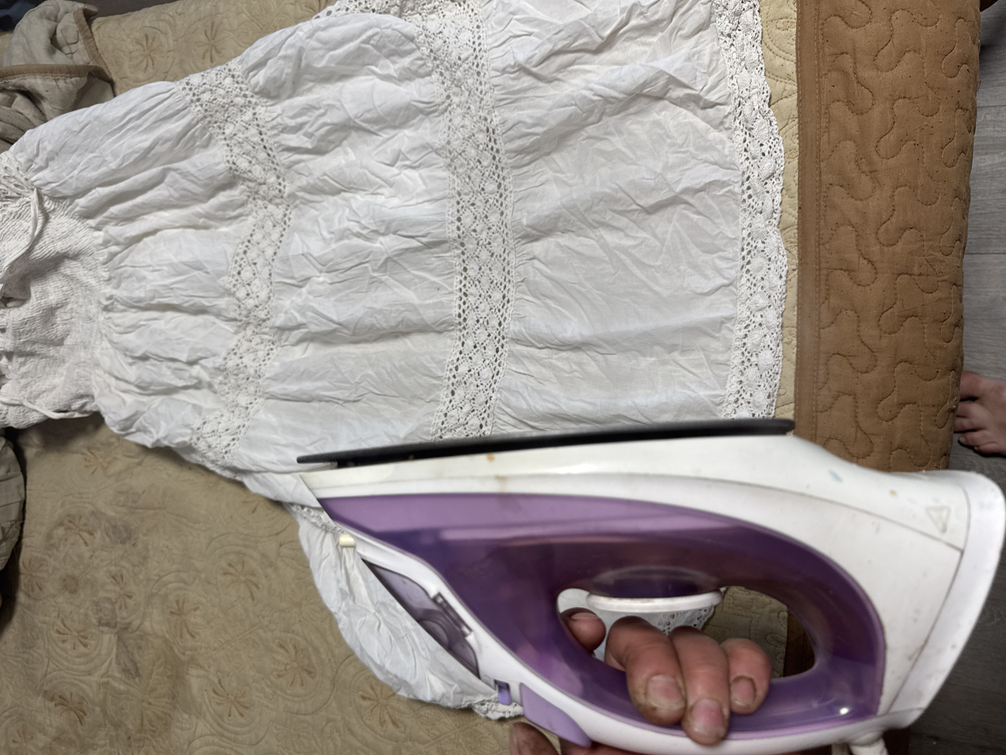 A white, lace-trimmed dress lies on a tan patterned bedspread. A white and purple iron is held by a hand with dirty fingernails, apparently being used to iron the dress. A section of brown quilted fabric is visible near the dress. The location is suspected to be within the city of Chaikovsky, Russia.
