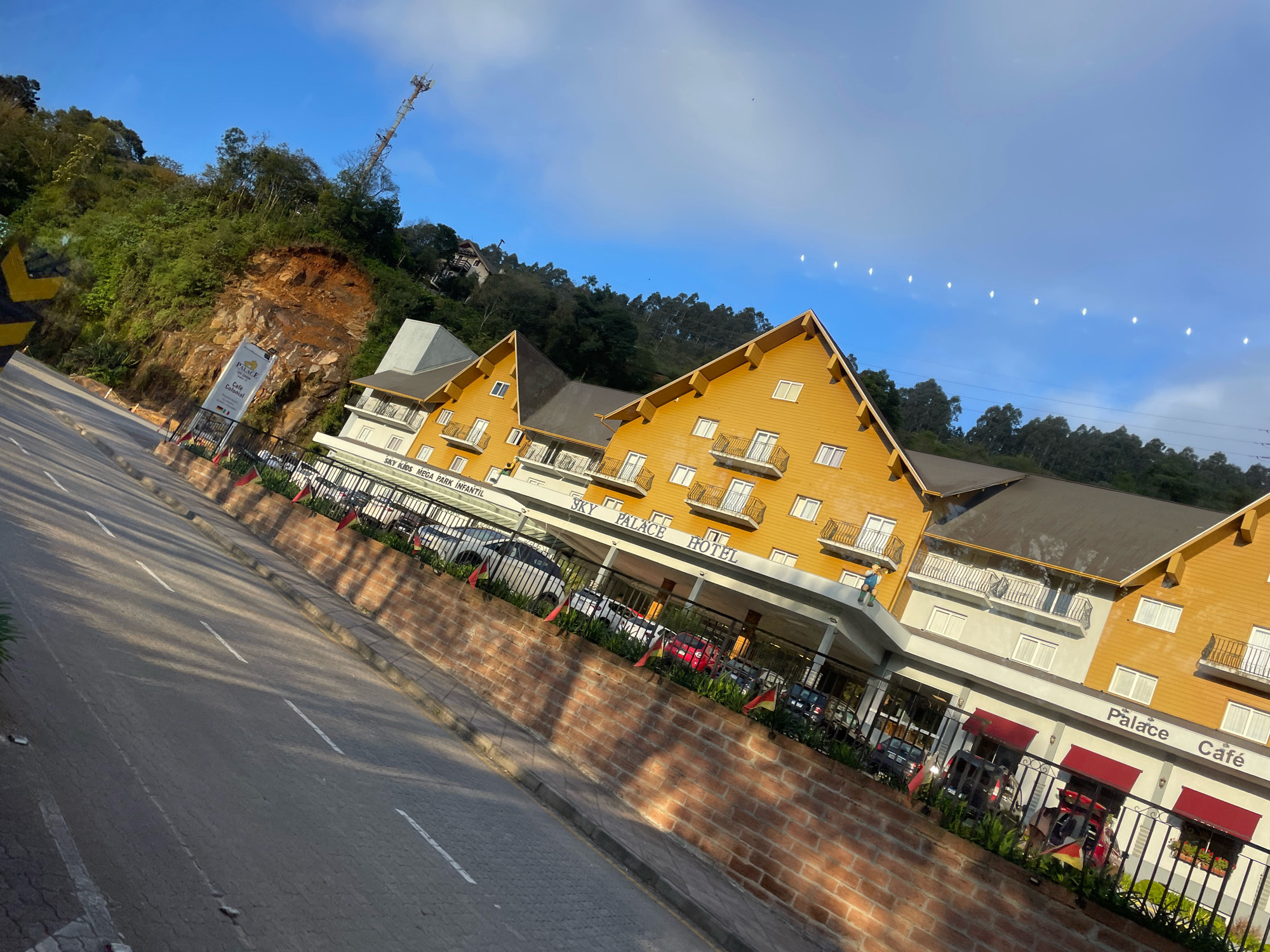 In the city of Gramado, Brazil, a hotel named "Sky Palace Hotel" is prominently featured. The hotel has a distinctive yellow facade with dark brown roofs, and multiple balconies with black railings. The entrance area is covered by a white awning, and the words "Palace Café" are visible above red awnings. The hotel is situated on a hillside, with lush green trees and a steep, rocky incline behind it. A telecommunications tower is visible on the hilltop. A paved road with white lane markings runs in front of the hotel, with a brick wall bordering the property. Several cars are parked in front of the hotel entrance. The sky is a clear blue with some white clouds and several small, bright spots which may be reflections or light flares. The image is angled, giving a dynamic perspective of the scene.
