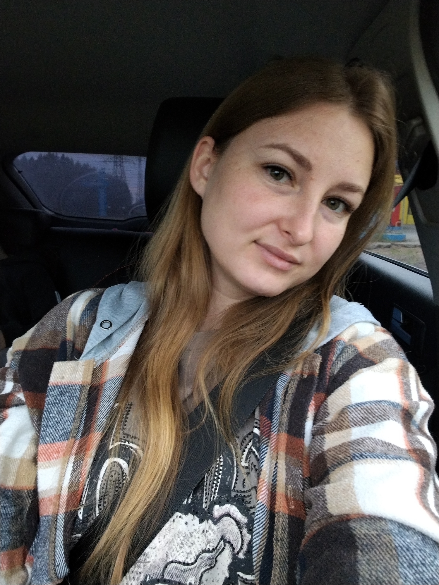 The image shows a woman in a car, wearing a seatbelt, a plaid shirt over a patterned top, and a hoodie. She has fair skin, light brown hair, and green eyes. The background includes a car window with a partial view of what seems to be a power line structure and trees outside, with what looks like colorful playground equipment visible on the right side. The image seems to have been taken inside the car. There are no details to suggest any information related to Noviy, Russia.