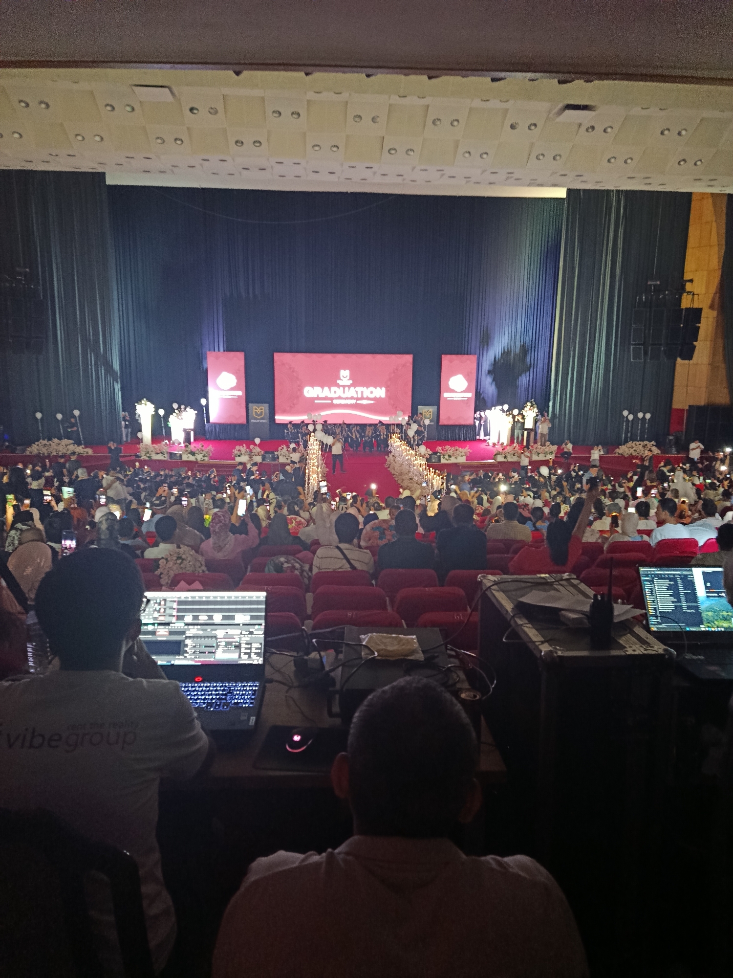 The image depicts a graduation ceremony taking place in a large, dimly lit auditorium. In the foreground, two individuals are seated, facing away from the camera, engrossed in operating technical equipment, likely for audio or visual production. One of them wears a t-shirt with the "vibe group" logo. In the background, on a raised stage decorated with flowers and lights, a group of people, presumably graduates and faculty, are visible. Two large screens display the word "GRADUATION" and a university logo. The audience is seated in rows of red chairs, many holding up mobile phones, suggesting they are documenting the event. The overall ambiance is one of celebration and formal occasion. The graduation ceremony is likely held in the Palace of International Forums in Tashkent, Uzbekistan, given its grandeur and typical use for such large-scale events.