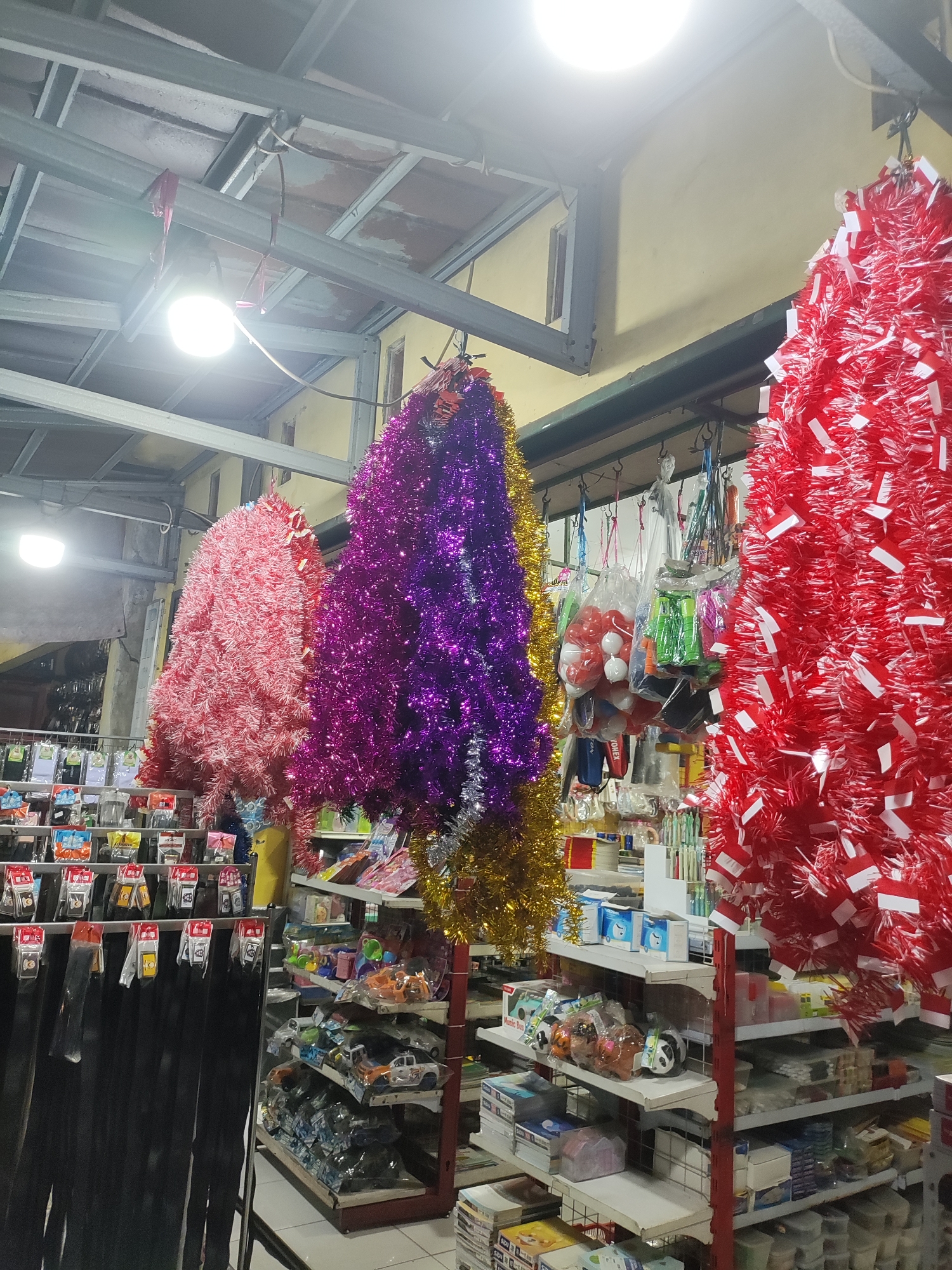 A low-angle, medium shot captures the interior of a brightly lit store in Wanajaya, Indonesia, filled with various merchandise. Dominating the foreground are three large, conical decorations resembling Christmas trees, made of vibrant tinsel in red, purple, and pink. These hang from the ceiling, suspended by thin strings.
Behind these festive decorations, shelves are stacked high with a diverse array of items. On the left, dark, long, thin objects hang in rows, possibly belts or fabric rolls. Above them, smaller items are displayed in packaging, some featuring red and yellow accents.
The middle section of the store reveals more shelving units. One shelf displays several plastic bags filled with what appear to be small balls, some white, some red, and other items in green plastic packaging. Below this, shelves are filled with various packaged goods, including boxes of tissues, toys such as toy cars and plush animals, and other unidentifiable products.
The background shows more shelving and a glimpse of what might be the store's exterior through a window or opening on the far left, suggesting it is a street-level establishment. The ceiling is a metallic structure with exposed beams and panels, and a bright, white light fixture hangs from the beams, illuminating the scene. The walls appear to be painted a pale yellow. The overall impression is that of a well-stocked general store or a shop specializing in party supplies and small goods.