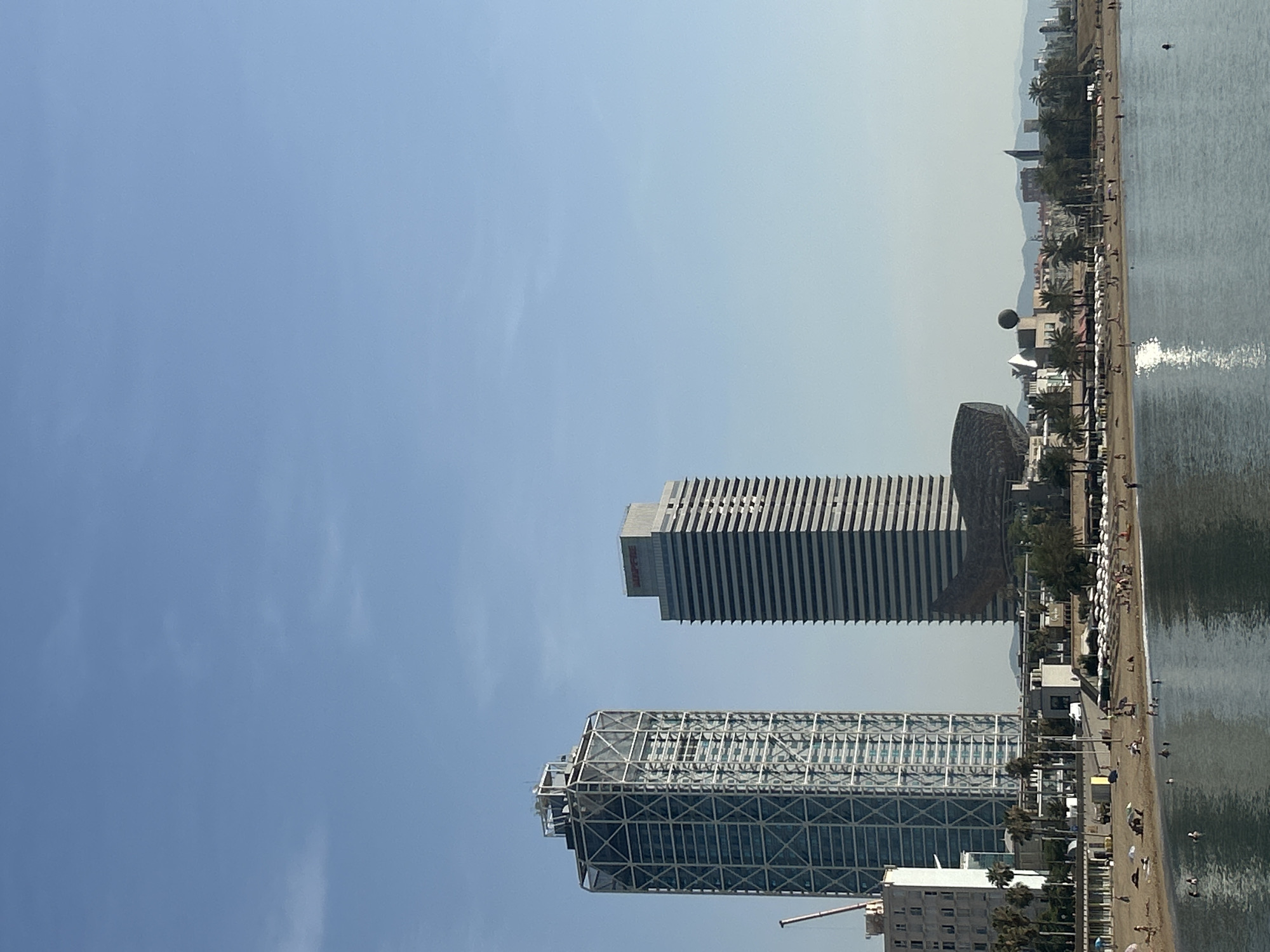 The image depicts a sunny day at a beach in Barcelona, Spain, with the iconic skyline of the city in the background. In the foreground, the calm blue water of the Mediterranean Sea reflects the bright sky and the buildings. The beach itself is populated with people sunbathing, swimming, and relaxing on the sand.
Dominating the skyline are two prominent skyscrapers. The one on the left is a modern, glass-clad building with a distinctive external lattice structure. To its right stands another tall building, characterized by a facade of horizontal gray bands, topped with a red logo. Between these two structures and further back, a unique, dark-colored building with a curved, almost organic shape is visible, resembling a fish or a wave.
A palm-lined promenade runs along the back of the beach, with rows of white umbrellas providing shade for beachgoers. A spherical hot air balloon or blimp is suspended in the sky behind the buildings, adding an unusual element to the scene. The horizon is further defined by a range of distant hills and the outlines of other buildings, suggesting the extensive urban sprawl of Barcelona. The overall impression is one of a vibrant, active beach resort on a clear, pleasant day.