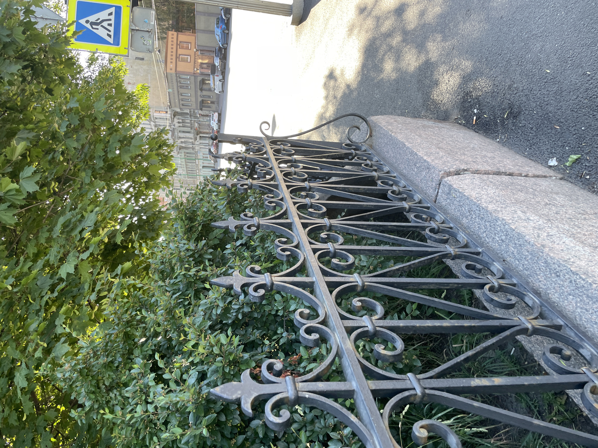 The image depicts a street scene in Saint Petersburg, Russia. In the foreground, a decorative black metal fence runs along a granite curb. Behind the fence, lush green bushes and tree leaves are visible, with sunlight filtering through the foliage. To the right, a paved sidewalk with shadows cast by overhead trees is present. Further down the street, a pedestrian crossing sign is mounted on a pole, indicating a crosswalk ahead. Buildings with architectural details line the street in the background, and several cars are parked or driving on the road. The overall impression is of a well-maintained urban environment on a sunny day.