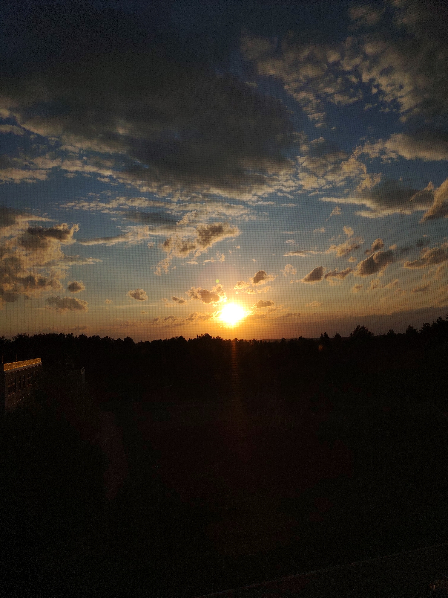 The image depicts a sunset viewed through a screen, possibly a window screen. The sun is the central focus, positioned near the horizon. The sky is filled with clouds, some dark and ominous, others lighter and reflecting the orange and yellow hues of the sunset. Below the horizon is a silhouetted landscape of trees and possibly buildings, making the landscape appear dark. The bottom portion of the image is obscured by shadow, suggesting a foreground closer to the viewer. The image appears to have been taken in or near Марковский, Russia, but without additional information, specific details about the location within the city cannot be determined. No people are visible.