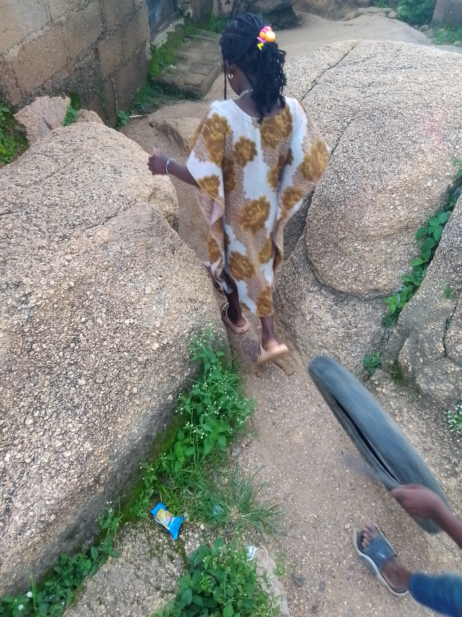 A rear view of a young Black girl with braided hair tied with a colorful hair accessory walks on a dusty path between large, uneven rocks. She wears a light-colored, loosely fitting garment with a brown and white pattern. Her right hand is extended as if reaching for something. To her right, a bicycle tire is being rolled along the path by an unseen person, whose arm and sandal are visible in the lower right corner of the image. Green vegetation and a discarded blue snack wrapper are present on the ground. The scene appears to be in a rocky, outdoor environment, likely on the outskirts of Jos, Nigeria, characterized by the natural rock formations and a rough, unpaved path.