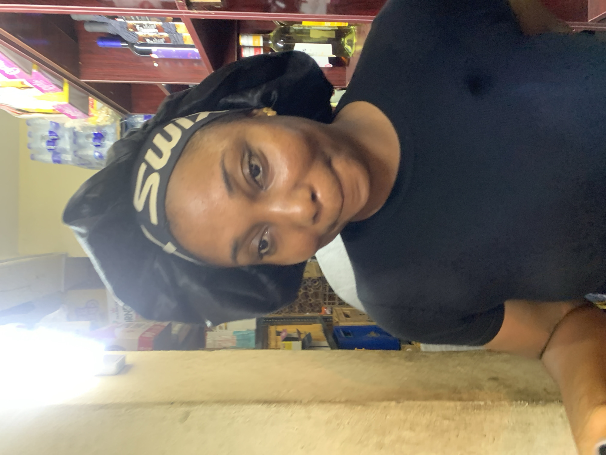 The image shows a medium close-up of a Black woman wearing a black t-shirt and a black bonnet with a white logo band. She is positioned in the foreground, facing slightly to the right. Her facial expression is ambiguous, with a slight smile and pursed lips. Behind her, shelves are visible filled with various items, including bottles of alcohol, water bottles, and boxed goods. The background suggests an indoor environment, likely a shop or store. The lighting appears to be artificial, with a bright light source on the left side of the frame casting a glow. The overall impression is that of a candid shot taken within a commercial setting.