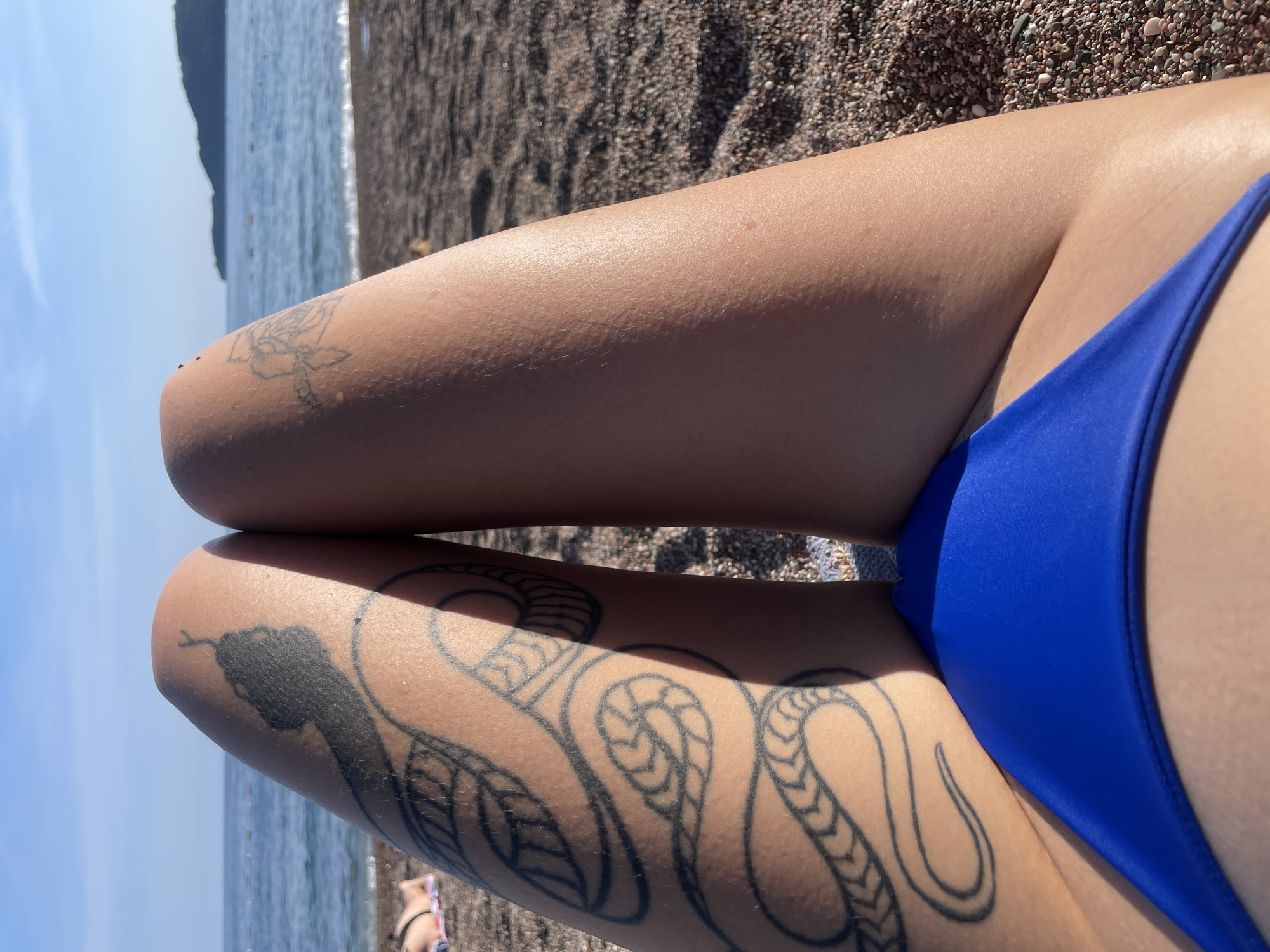 A close-up, slightly angled shot shows a person's crossed legs at a beach in Rafailovići, Montenegro. The person is wearing a blue swimsuit bottom. On the lower leg, there is a detailed tattoo of a snake winding around the limb, with the snake's head and leaves visible. On the upper leg, near the knee, there's a smaller, less detailed tattoo. The legs are positioned against a backdrop of the beach, with the sand visible, and the sea and sky in the distance. An island is barely visible on the top left of the image.