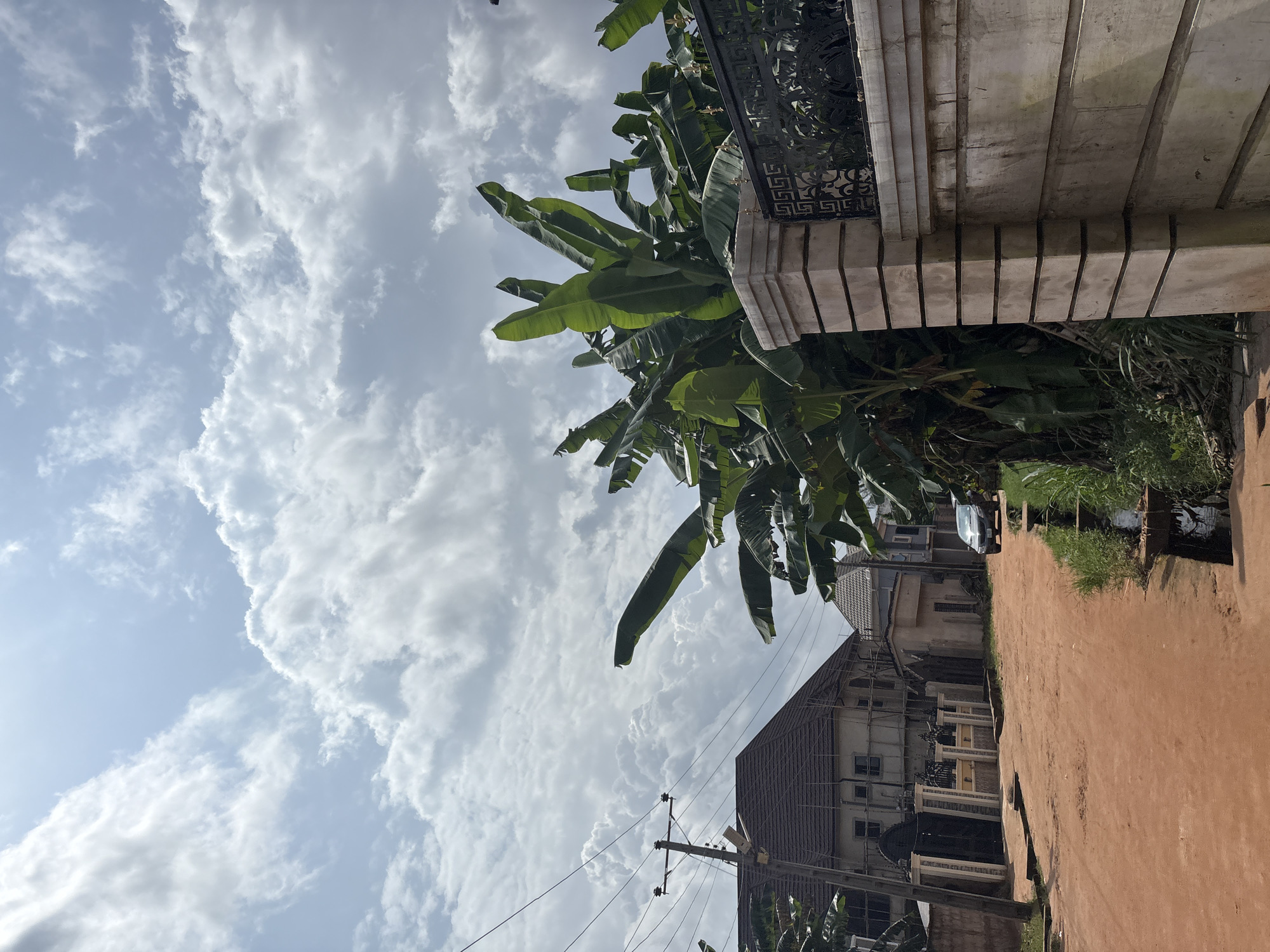 A dirt road in Obosi, Nigeria, leads towards a building under construction, with scaffolding visible. A utility pole with electrical lines stands to the left. Banana trees line the right side of the road, behind a concrete wall with a decorative metal fence. A dark-colored sedan is parked on the right side of the road. The sky is filled with dramatic, white, cumulus clouds.