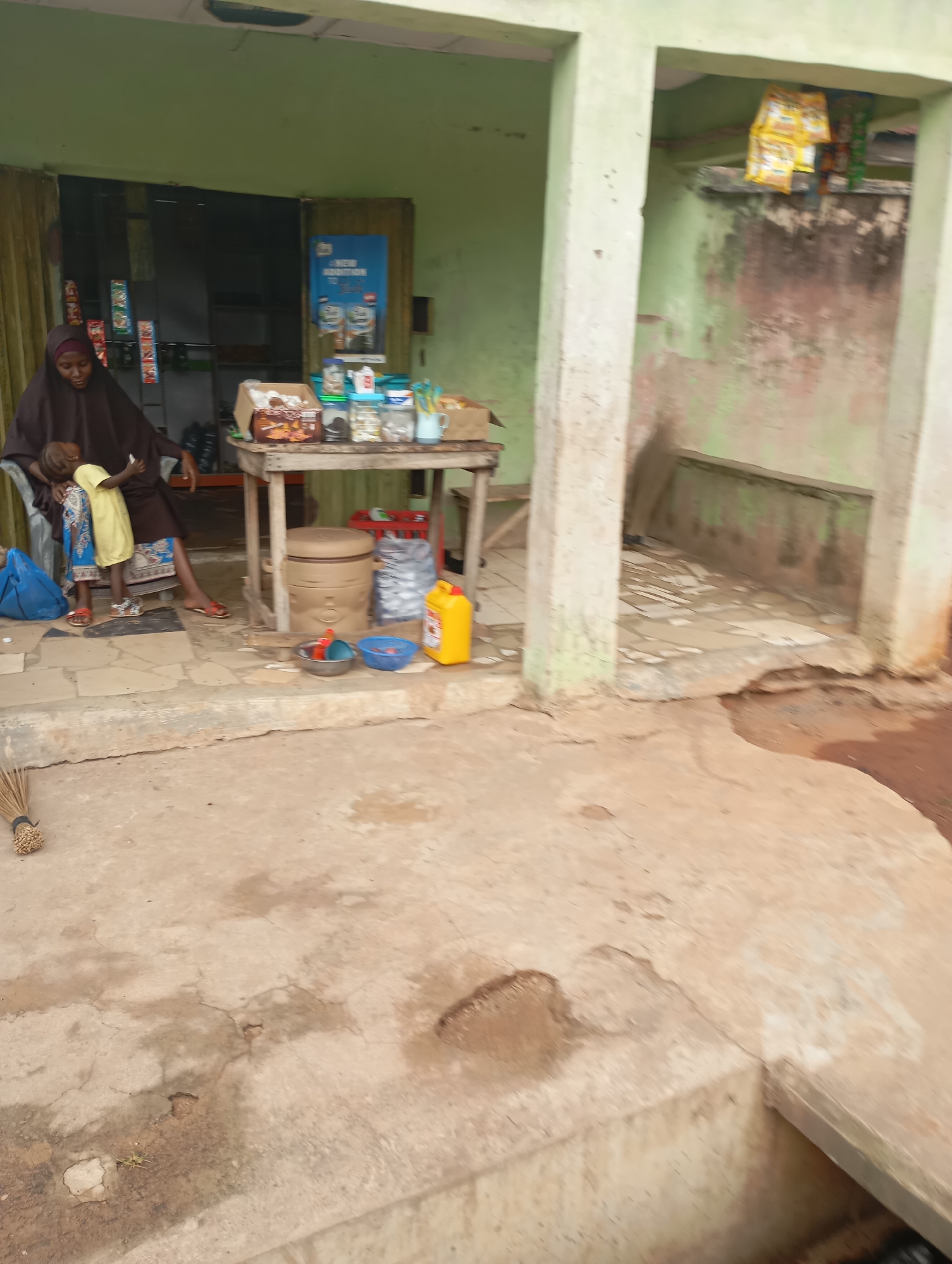 In Shendam, Nigeria, a woman wearing a brown hijab and abaya is seated, holding a sleeping child. To her right, a young child stands, looking towards her. They are positioned on a tiled concrete porch in front of a shop with a green, faded exterior. The shop's entrance is open, revealing shelves stocked with various goods, including what appear to be packaged snacks. A wooden table in front of the shop displays more items, such as jars and boxes. To the right of the woman, a large, brown, cylindrical container rests on the ground, accompanied by a yellow plastic jerrycan and two small bowls. Further to the right, a section of the building features a light green wall and two concrete pillars. The foreground consists of a cracked, sandy concrete ground, with a broom leaning against a low wall.