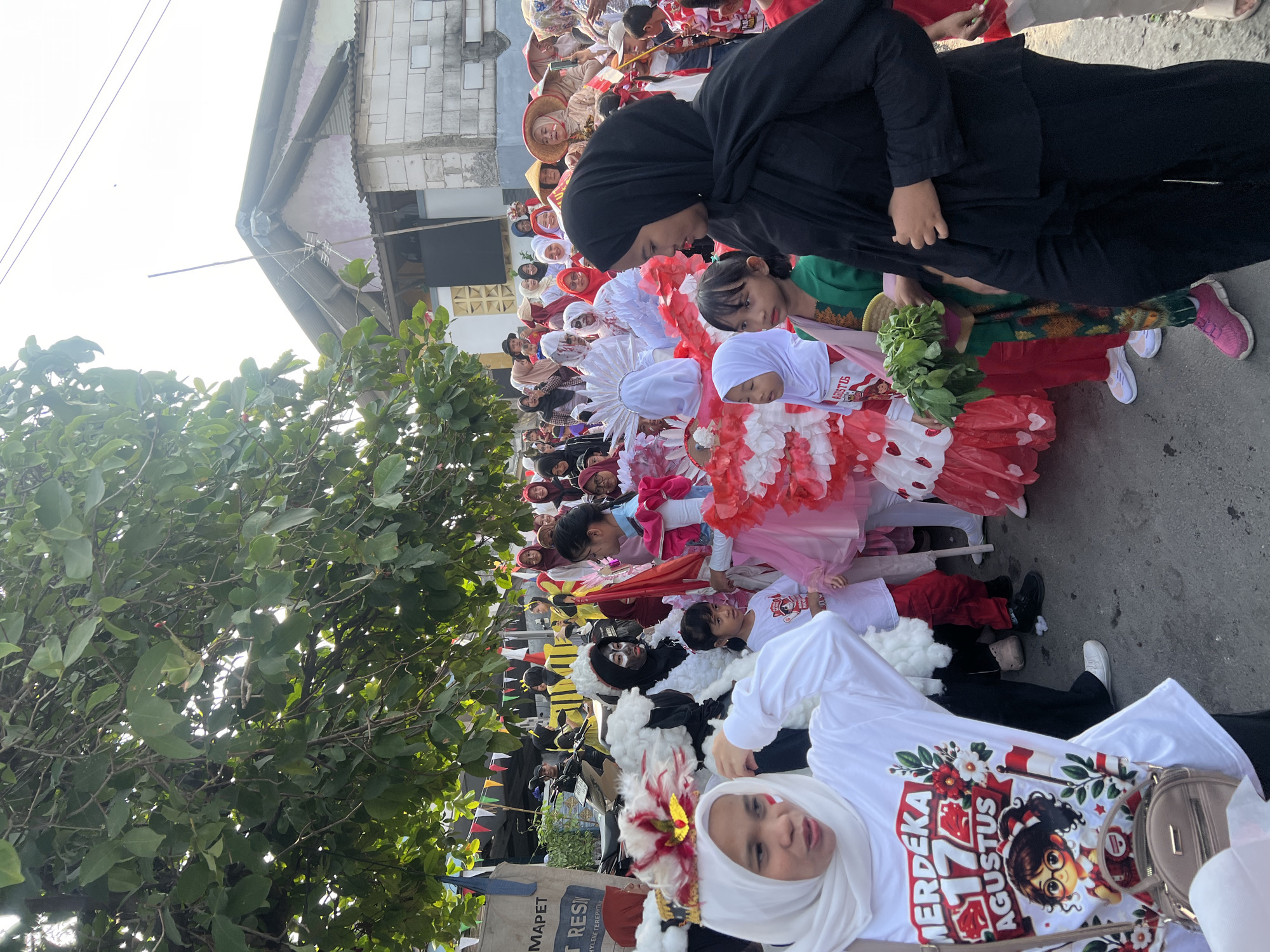 In Klapanunggal, Indonesia, a parade is taking place on a street lined with trees and buildings. A group of children and adults are participating, many dressed in traditional Indonesian attire or costumes. Some children wear white shirts with "MERDEKA 17 AGUSTUS" printed on them, signifying Indonesian Independence Day. Others are dressed in elaborate red and white outfits, and some have face paint. People are walking and interacting, suggesting a festive atmosphere. The environment appears to be a residential or semi-urban area, with buildings and a clear sky visible.