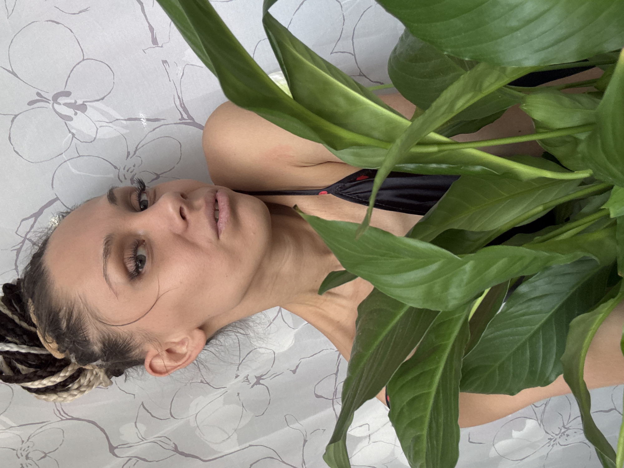 A woman with braided hair poses with a large green plant. She is wearing a black top and is lying on a white and gray floral patterned surface. The location is in the city of Unknown, Unknown.