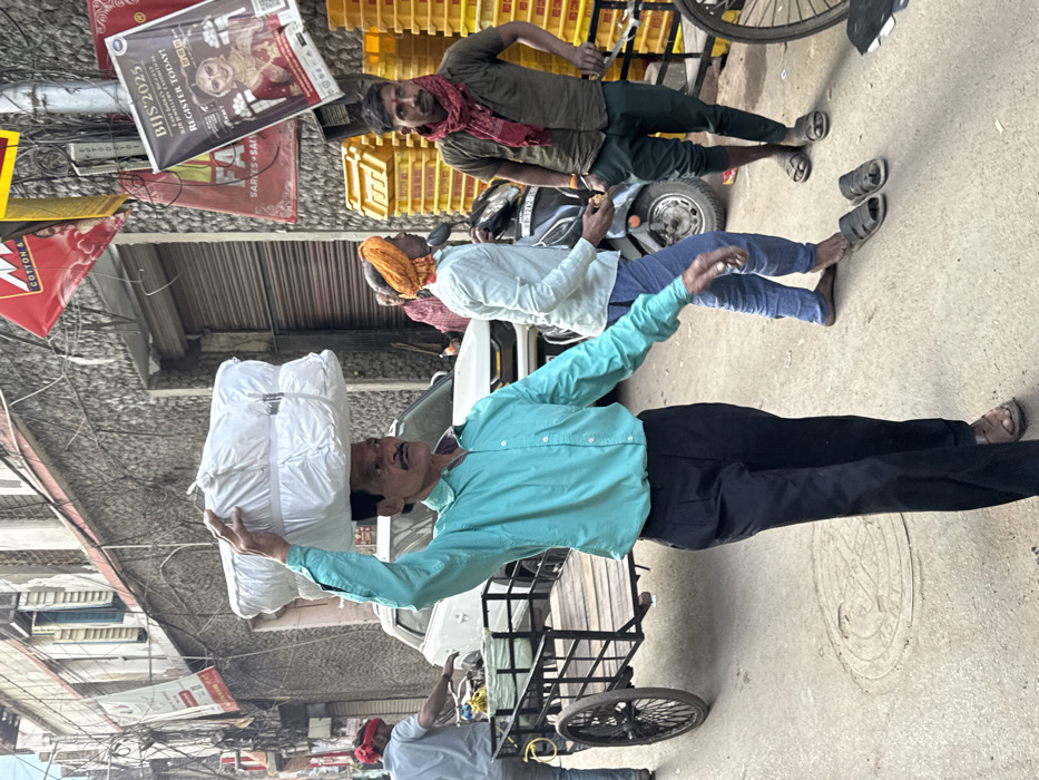 This image captures a daylight scene on a bustling commercial street in Kolkata, India.
In the foreground, an adult male, approximately 40-50 years old, with a mustache, wearing a light blue long-sleeved button-down shirt, dark trousers, and brown sandals, is walking towards the right side of the frame. He is balancing a large, rectangular, white, plastic-wrapped bundle on his head, supporting it with his raised right hand. A circular metal manhole cover is visible on the concrete ground near his feet.
In the mid-ground, from left to right: an adult male, partially visible, wearing a light shirt and red head covering, pushes a black metal hand-pulled cart with wooden slats and two spoked wheels. Behind him, a white SUV is parked or moving slowly. To the right, another adult male, wearing a light-colored long-sleeved shirt and blue pants, with an orange turban, stands facing left, seemingly having just removed his two dark sandals which lie on the ground in front of him. He is holding a small, light-colored object in his right hand. A black scooter is parked directly behind him. To his right, an adult male with a thin mustache, wearing an olive green short-sleeved shirt, dark green pants, and a red scarf around his neck, stands with his right hand on his hip, looking towards the viewer.
The background consists of multi-story buildings with textured, likely stone or concrete, facades. Numerous exposed electrical wires crisscross overhead. Multiple commercial banners and posters are affixed to the buildings. A prominent poster on the right advertises "BIJS 2025" (Bengal International Jewellery Show), featuring a woman in traditional bridal attire and text encouraging registration. Other banners partially visible include "COTTON &" and "SARIES". A stack of bright yellow plastic crates is visible on the right side of the frame, behind the man with the red scarf. The overall environment suggests a vibrant commercial thoroughfare.