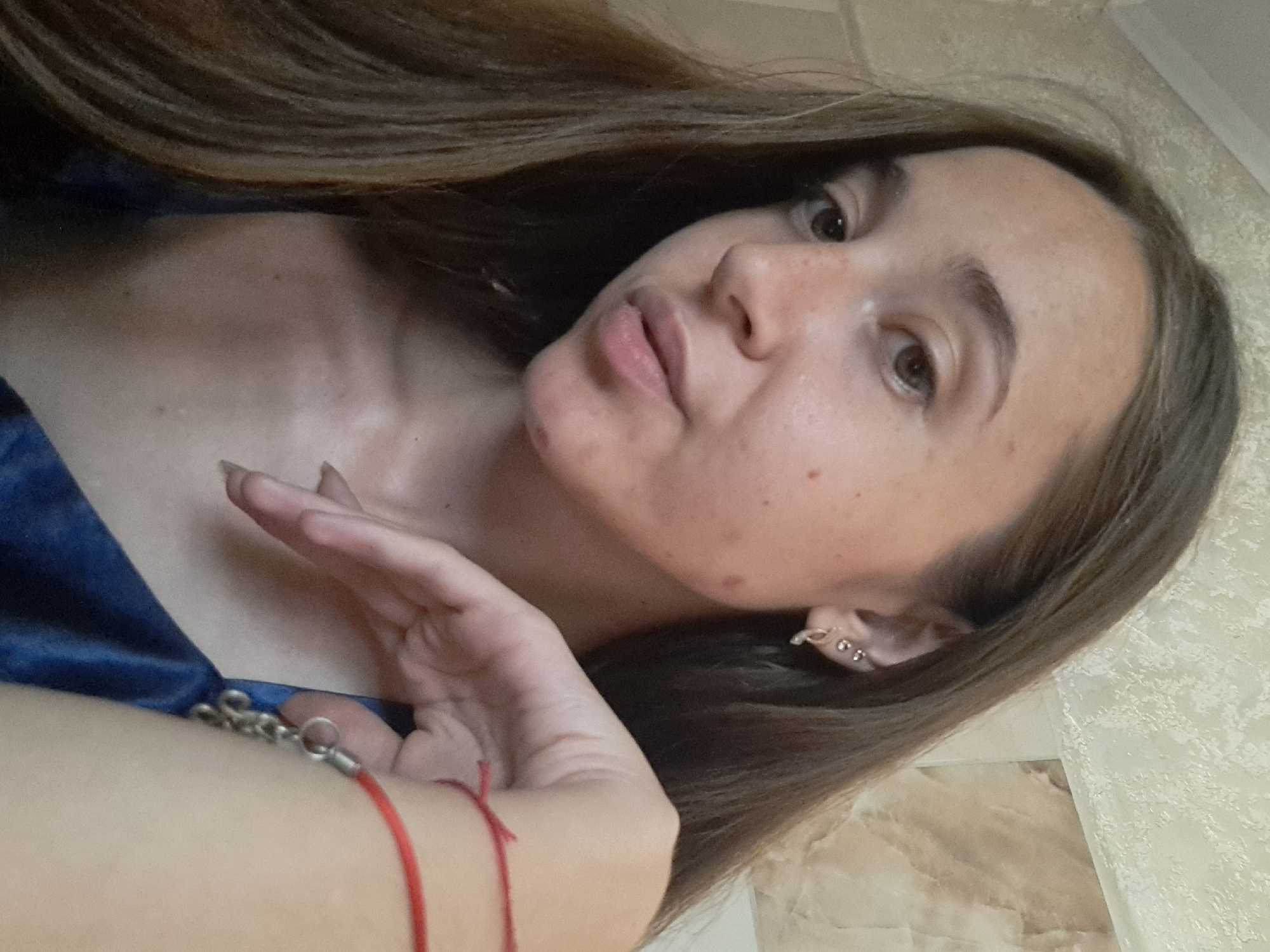 A close-up, waist-up shot shows a young woman with long, dark brown hair, facing the camera at a slight angle. Her skin has freckles and some visible blemishes. She is wearing a dark blue top. Her left hand rests on her chest, adorned with a red string bracelet and a silver charm bracelet. The background appears to be an indoor setting with textured wallpaper in muted tones. The lighting is soft, casting a warm glow on her face.