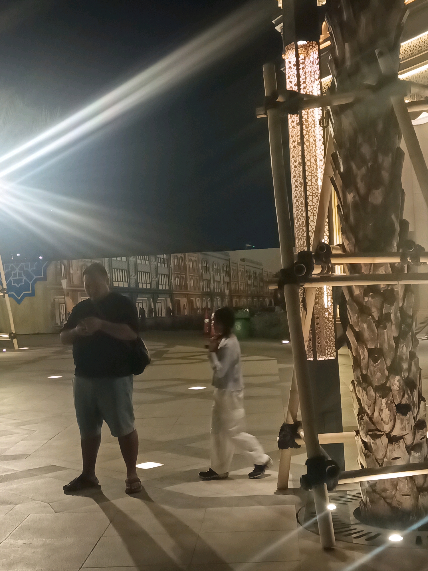 The image depicts a nighttime scene in Ancol, North Jakarta, Indonesia, with two individuals and architectural elements in the foreground. On the left, a man wearing a dark t-shirt and light-colored shorts stands with his back to the camera, holding a dark bag over his shoulder and looking down at his hands. To his right and slightly in front, a person in light-colored clothing walks away from the camera, their body angled to the left. In the immediate foreground, a large, textured tree trunk is partially visible, supported by wooden scaffolding and illuminated by an ornate, patterned light fixture within the scaffolding. The ground appears to be paved with light-colored tiles and features circular lights embedded in the surface. In the background, a building with a repeating architectural pattern of windows and decorative elements is illuminated, suggesting an outdoor public space. Strong light flares from the left side of the image indicate a bright light source, possibly a spotlight or camera flash, creating lens flare effects.