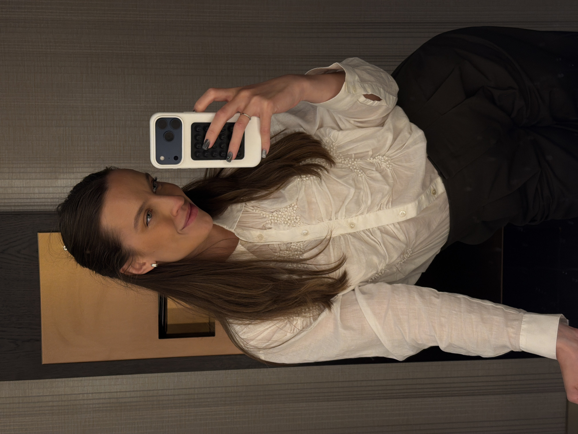 A woman is taking a selfie in a room with horizontal striped wallpaper. She is wearing a white long-sleeved blouse and black pants. She is holding a phone with a textured white case in her right hand. The background includes a gold-colored rectangular object. The city of Budva, Montenegro is not discernable in the image.