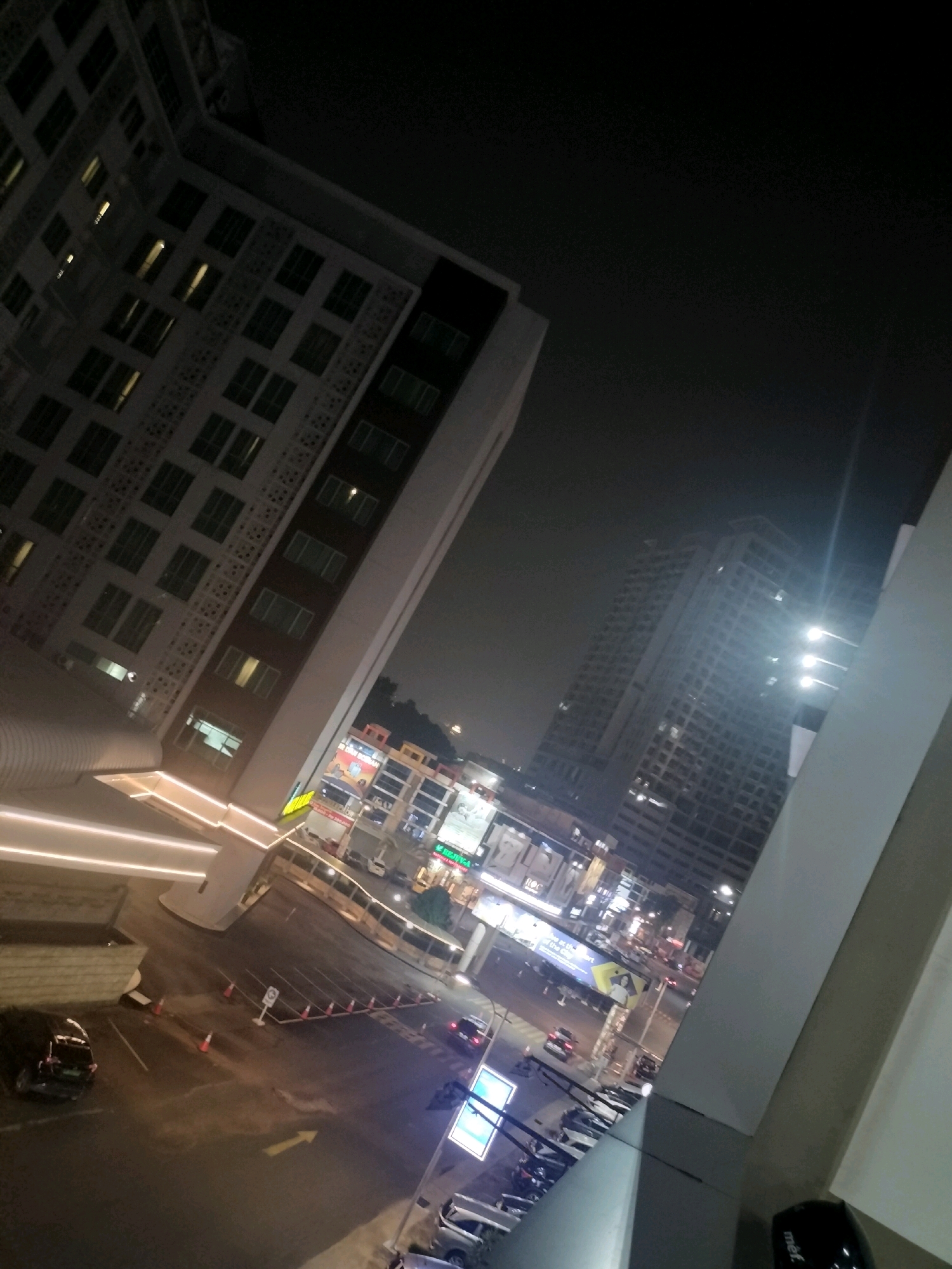 The image captures a nighttime urban scene, likely in Batam, Indonesia. A tall, modern building with numerous illuminated windows dominates the left side of the frame, its facade featuring a repeating geometric pattern. The building is angled, giving a sense of depth to the composition. In the background, other high-rise buildings are visible, their lights reflecting in the dark sky.
Below the main building, a street level view reveals a parking area with several cars, including a dark-colored sedan on the far left. Orange traffic cones are placed along the edge of the parking lot, marking a boundary or lane. A prominent yellow arrow is painted on the asphalt, indicating a direction. Further down the street, various commercial establishments are lit up, with signs and illuminated advertisements suggesting a bustling area. One sign displays the word "KENYA". A few vehicles are seen on the road, moving through the urban landscape. The overall atmosphere is one of a contemporary city at night, with artificial lights illuminating the structures and streets.