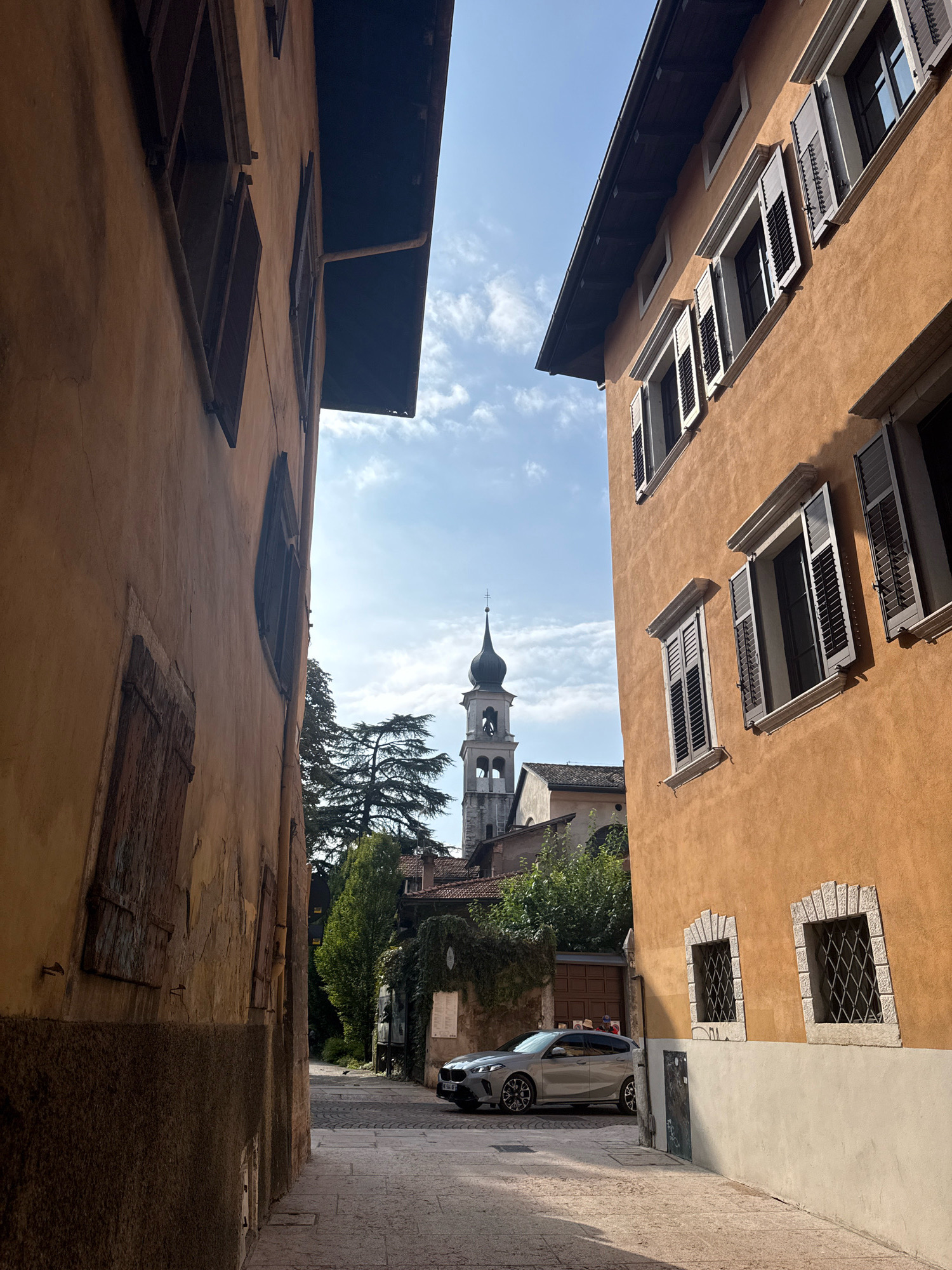 A narrow cobblestone street in Trento, Italy, is framed by tall, ochre-colored buildings with dark shutters. A silver hatchback car is parked on the right side of the street, facing away from the viewer. In the distance, a church bell tower with a black spire rises above the rooftops. Lush green trees are visible behind the buildings, and the sky is a clear blue with a few scattered clouds. The scene is bathed in bright sunlight, casting sharp shadows on the street and buildings.