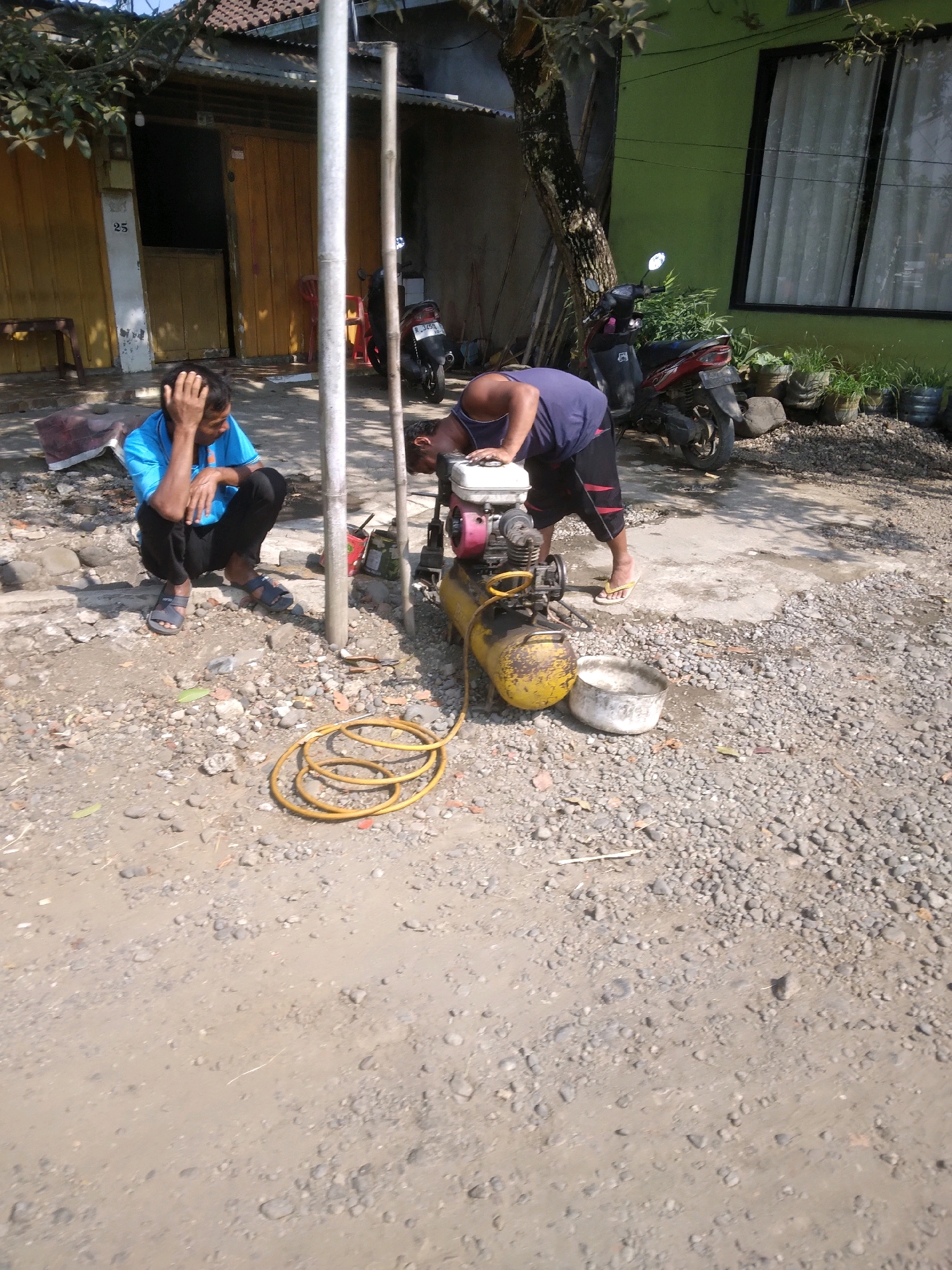 In the village of Desa Pamijen, Kecamatan Sokaraja, Indonesia, a man in a blue shirt and black pants is crouched on the ground, resting his head in his hand. Nearby, another man, wearing a dark purple shirt and dark shorts, is leaning over a yellow air compressor with a red engine, appearing to be working on it. A coiled yellow hose lies on the gravelly ground in front of the compressor. In the background, a building with a yellow facade and a brown wooden door is visible, along with a tree and a green building. Motorcycles are parked in the background. The scene is set in an outdoor area with a dirt and gravel ground surface.