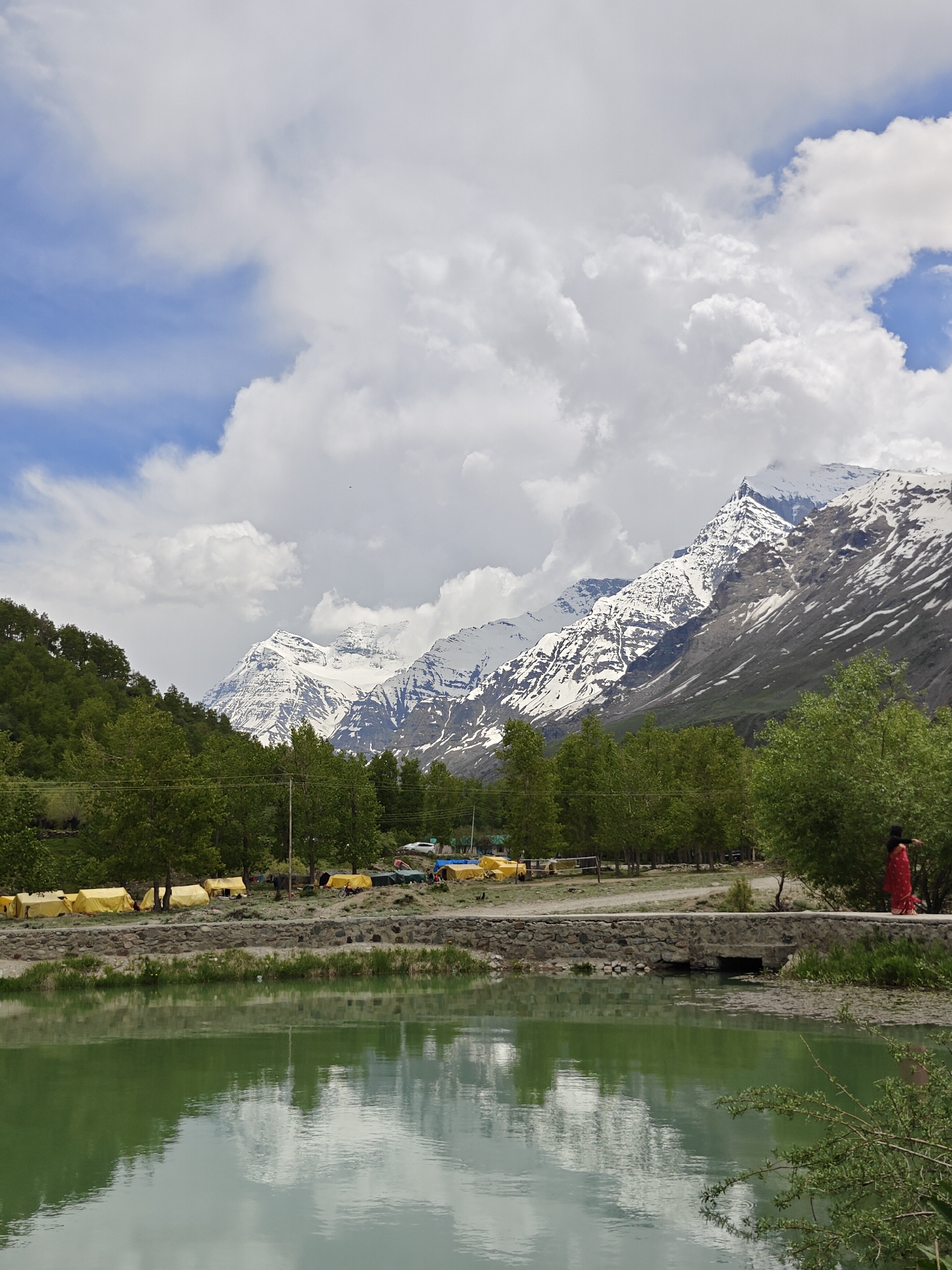 The image shows a landscape in Yamling, India. In the foreground is a body of water, its surface reflecting the sky and surrounding landscape. Behind the water is a strip of land with a low stone bridge. A person in a red garment walks along the pathway on the bridge. Behind the bridge is a line of trees. Yellow tents are interspersed among the trees. In the background, large snow-capped mountains rise, partially obscured by clouds. The sky is a mix of blue and white, with large, puffy clouds.