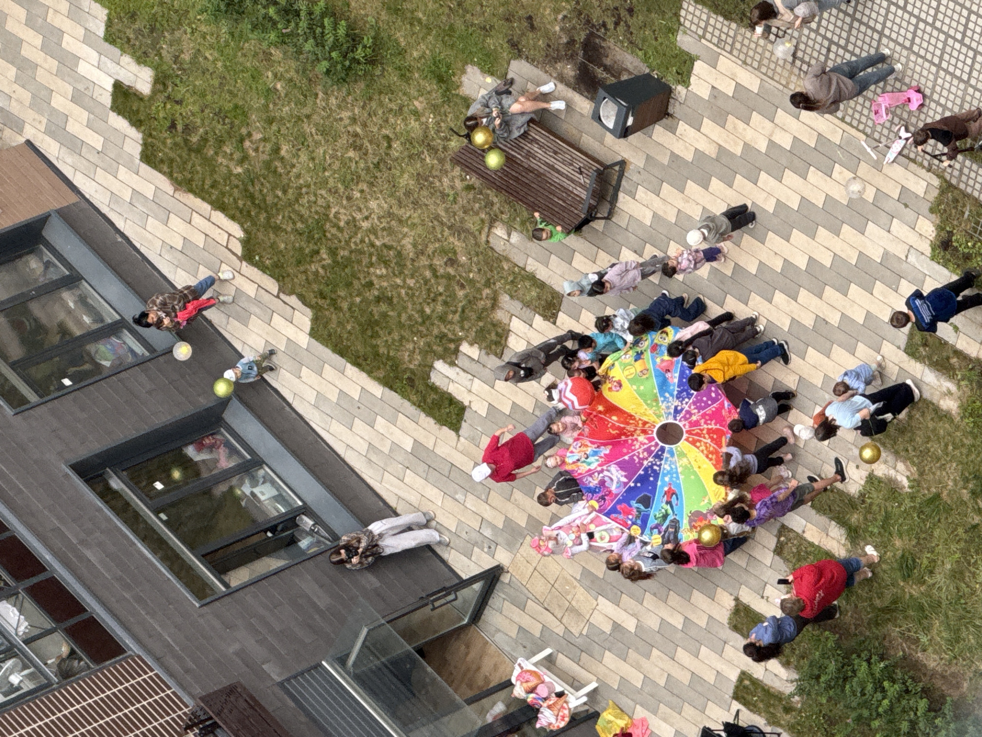 In Lyubertsy, Russia, a group of children and adults are gathered in an outdoor courtyard area. The setting appears to be near a modern residential building with dark brick facade and large windows. The ground is paved with light and dark rectangular tiles, interspersed with patches of green grass and a small tree.
In the center of the paved area, a large, colorful parachute game is spread out on the ground, with many children and a few adults surrounding it, actively participating. Several children are holding onto the edges of the parachute, and some appear to be laughing or engaged in conversation. One adult, wearing a red shirt and white cap, seems to be directing or observing the activity.
To the left of the parachute, there is a white table with several items on it that resemble ice cream cones or pastries. A woman in a patterned shirt is standing near a glass door, holding a camera and seemingly taking pictures of the event.
Further to the right, a wooden bench is positioned on the paved area. A woman in a gray coat is seated on the bench, looking at her phone, with two yellow balloons nearby. Adjacent to the bench is a dark gray trash receptacle.
Other individuals are scattered throughout the scene. Some adults are standing and observing the children, while others are walking. A few children are playing independently or are being held by adults. One child is seen with a yellow balloon, and another small child is near a pink toy scooter. The overall atmosphere suggests a celebratory or recreational gathering.
