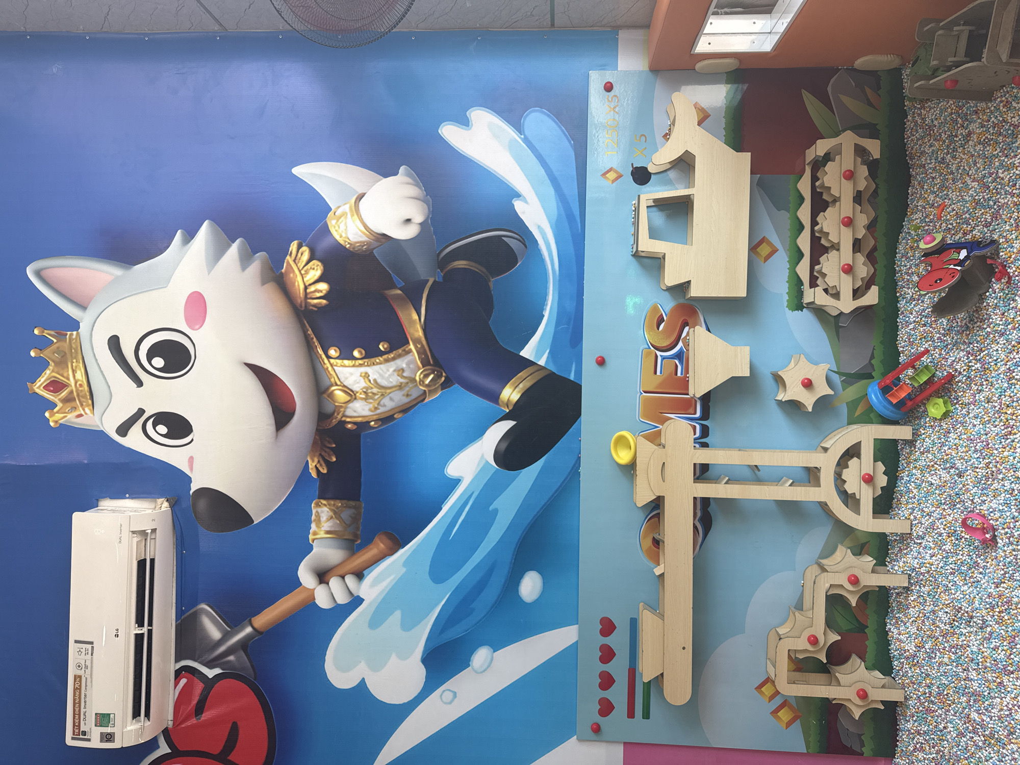 The image depicts a colorful indoor play area, likely a children's entertainment center. The background features a large mural of a cartoon wolf character wearing a crown and a blue and gold uniform, standing on blue waves. To the left of the mural, an LG brand air conditioner is mounted high on the wall. Below the mural, a wooden play structure is installed against the wall. This structure includes various interactive elements like a balance scale, gears, and ramps, all designed for children's engagement. The play area is filled with a soft, colorful ball pit. Several small toys are scattered within the ball pit, including a red and blue toy with wheels and a pink ring. The overall environment appears to be designed for children's recreation and amusement. The location is identified as Bac Nha Trang Ward, Vietnam.