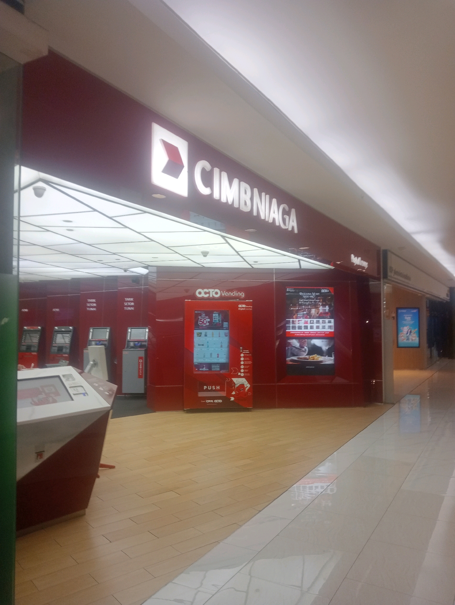 The image shows a bank branch of CIMB Niaga in South Jakarta, Indonesia. The bank features a prominent red facade with the CIMB Niaga logo. Several ATMs and a vending machine are visible, along with digital displays showing advertisements and information. The floor is a combination of wood and tile. The scene appears to be an indoor shopping mall or a bank lobby. There are no people clearly visible in the image.