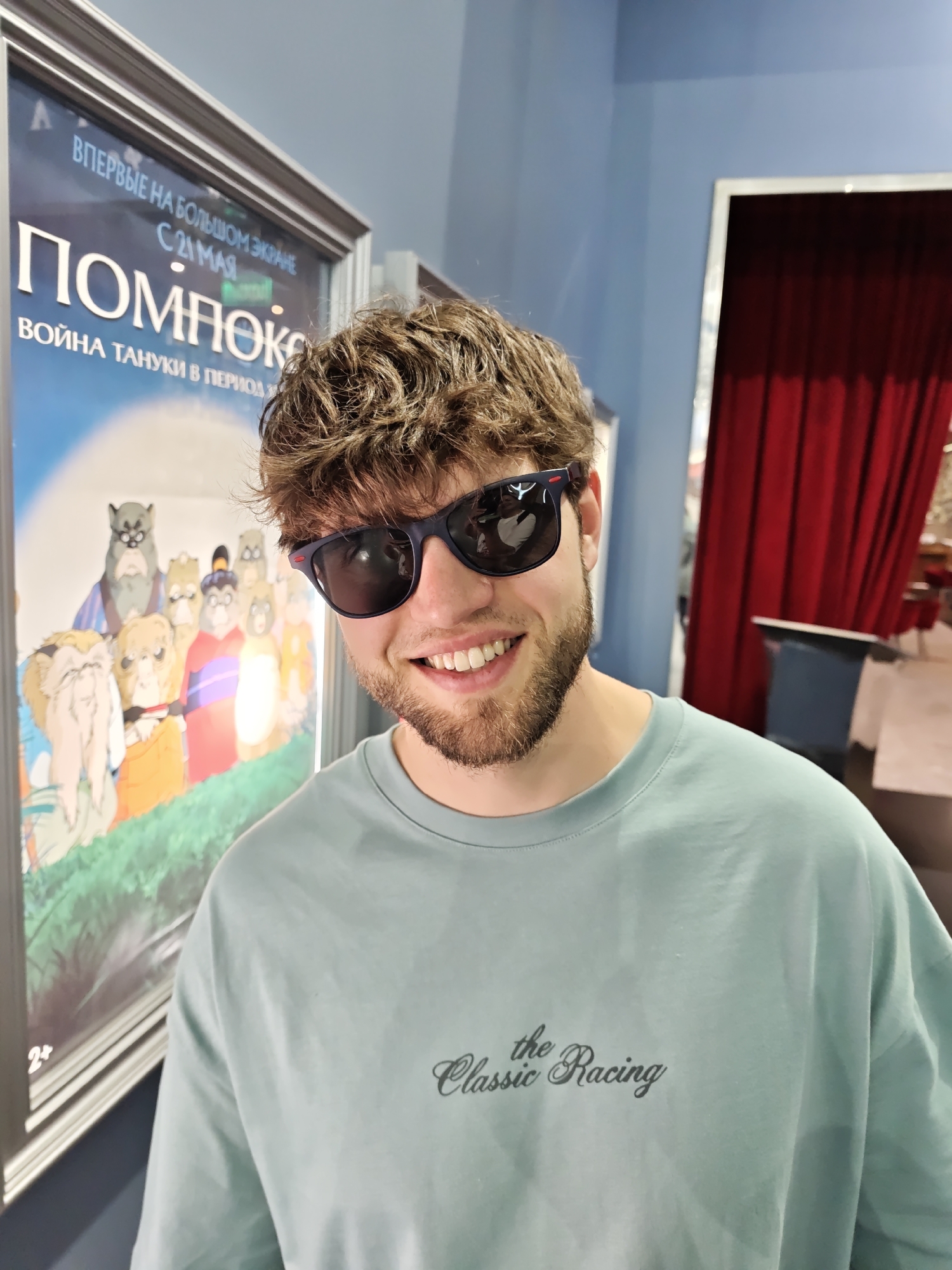 A young man with brown hair and a beard is smiling and wearing sunglasses in front of a poster. The poster promotes a film titled "Pompo-ke: The Tanuki War in the Period" and indicates its screening from May 21. The poster has the image of animated characters. The man is wearing a light blue t-shirt with the words "the Classic Racing" written on it. Behind him, there's a blue wall and a doorway with red curtains. The location is Novosibirsk, Russia, based on the Cyrillic text on the poster.
