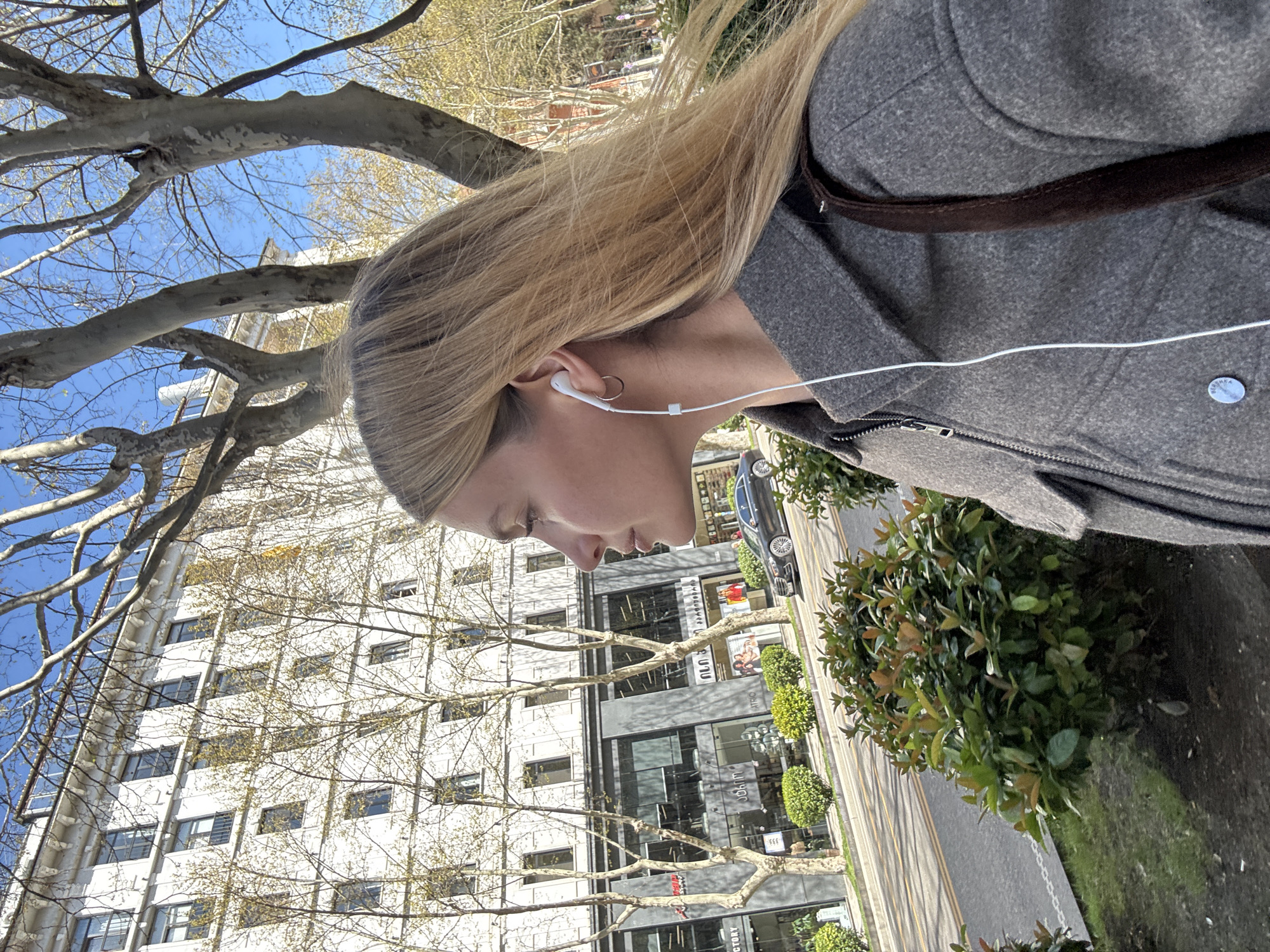 A woman with long blonde hair is wearing a gray coat and white wired earphones, standing on a city street. She is facing left in profile. Behind her is a multi-story white building with numerous windows. Trees with bare branches are interspersed between the woman and the building. Along the right side of the frame is a sidewalk with greenery. A road with a parked car is visible. The location is Tbilisi, Georgia, as determined by text on the building.